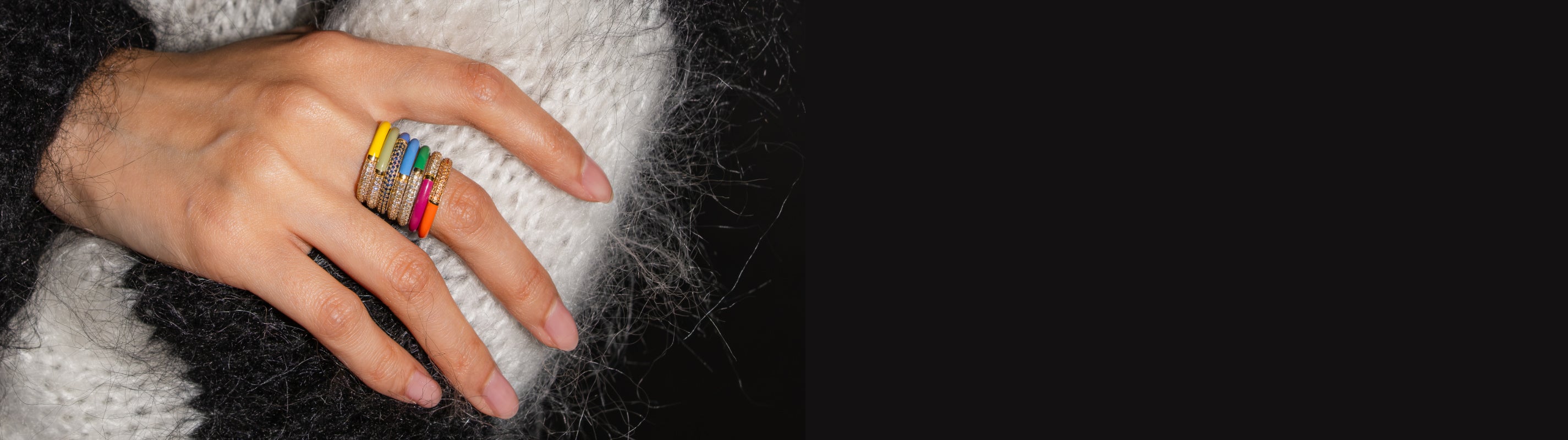A close-up of a hand wearing a stack of colorful rings, resting on a white knit sweater against a dark background.