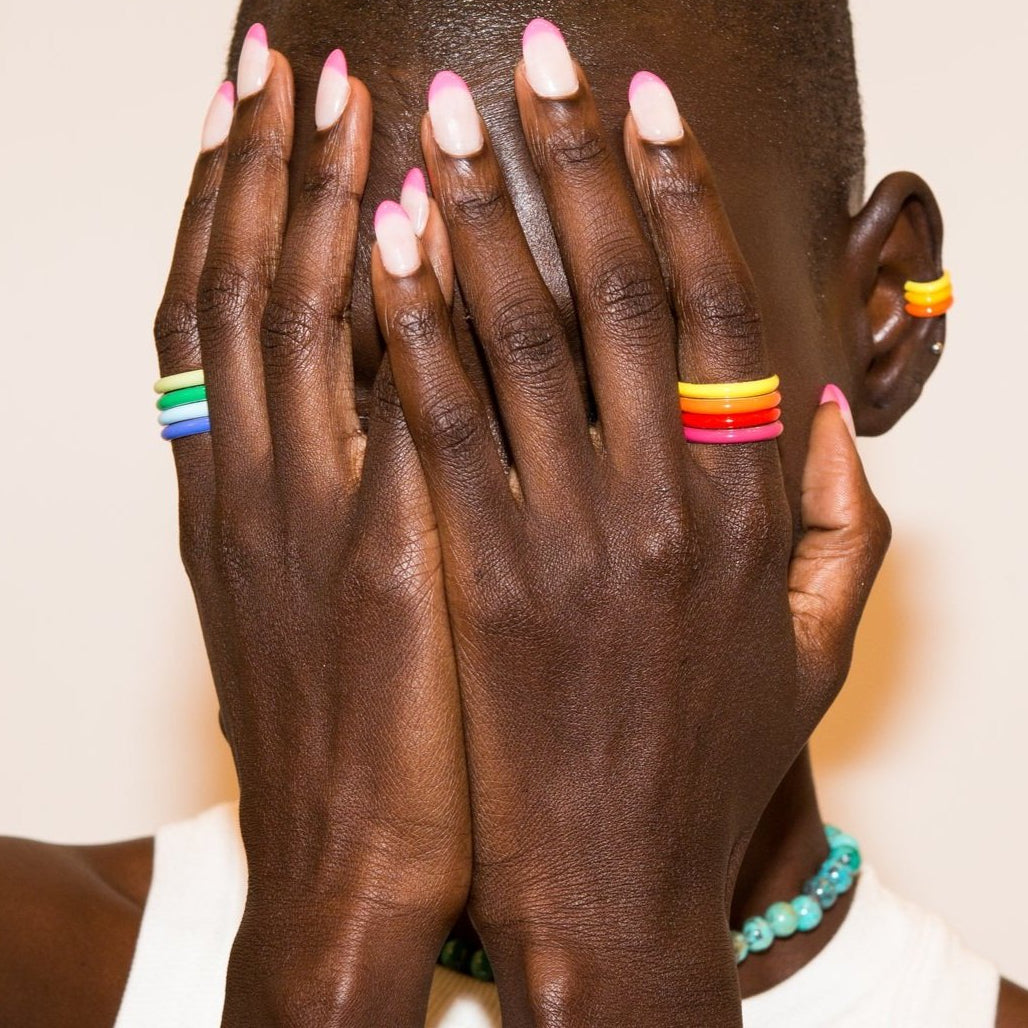 A close-up lifestyle shot of a person with dark skin, pink-tipped nails, and colorful stacked rings on their fingers and ear, covering their face with their hands against a plain light background.