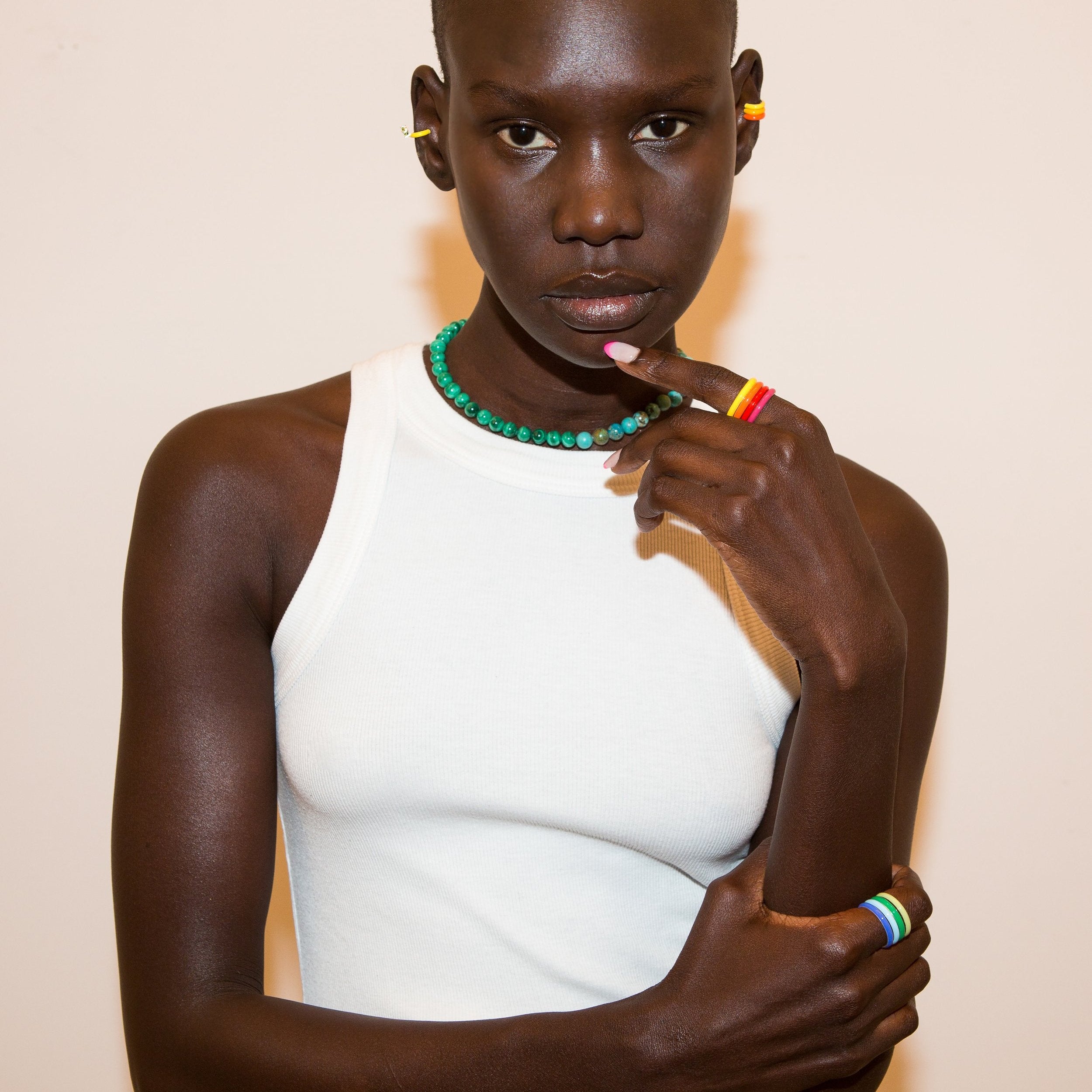 A model wearing a white ribbed tank top, a green beaded necklace, yellow hoop earrings, a rainbow ring, and a rainbow stack of eight rings, posed against a solid light beige background.