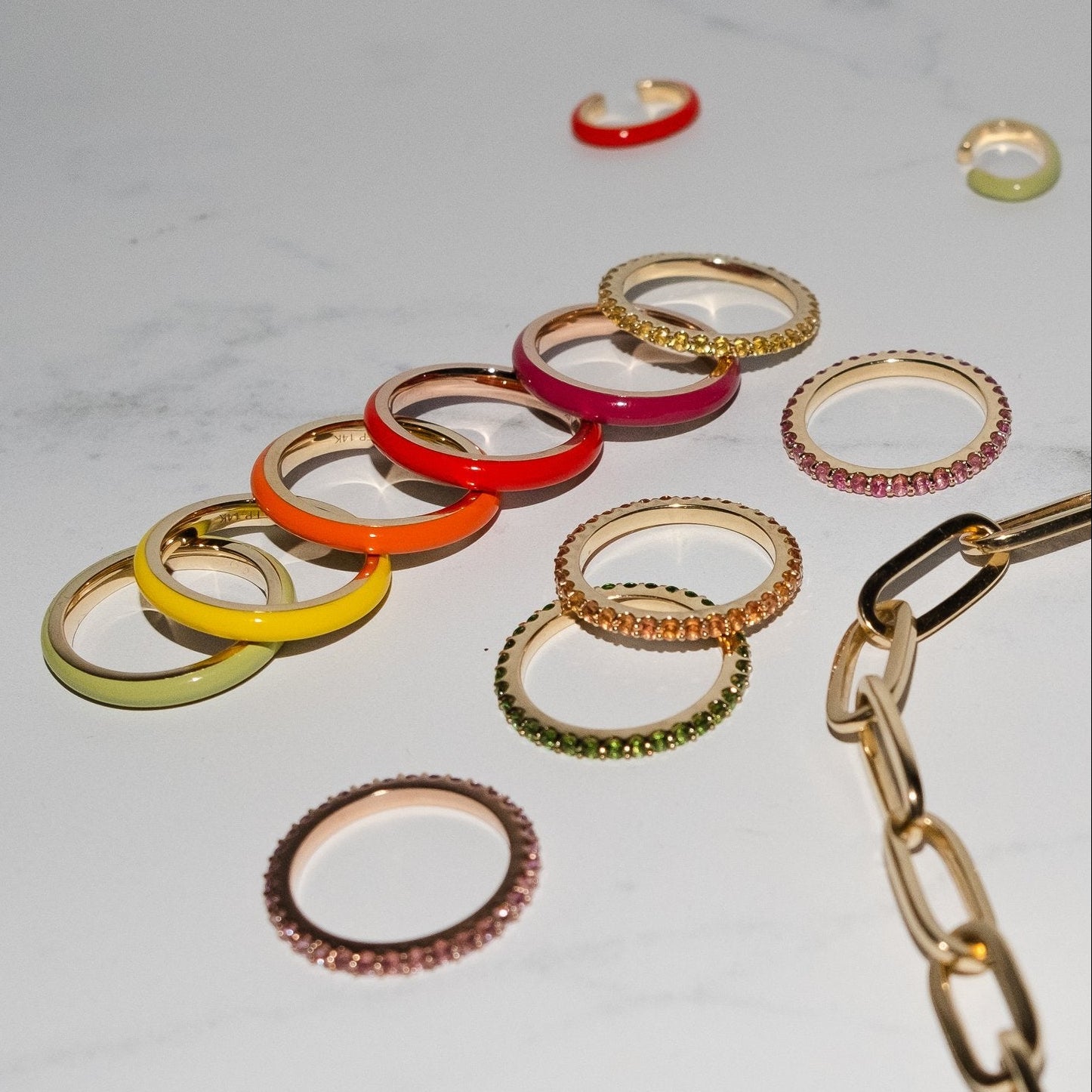A collection of colorful, thin band rings with gemstone rings, arranged on a light marble surface alongside a gold chain.