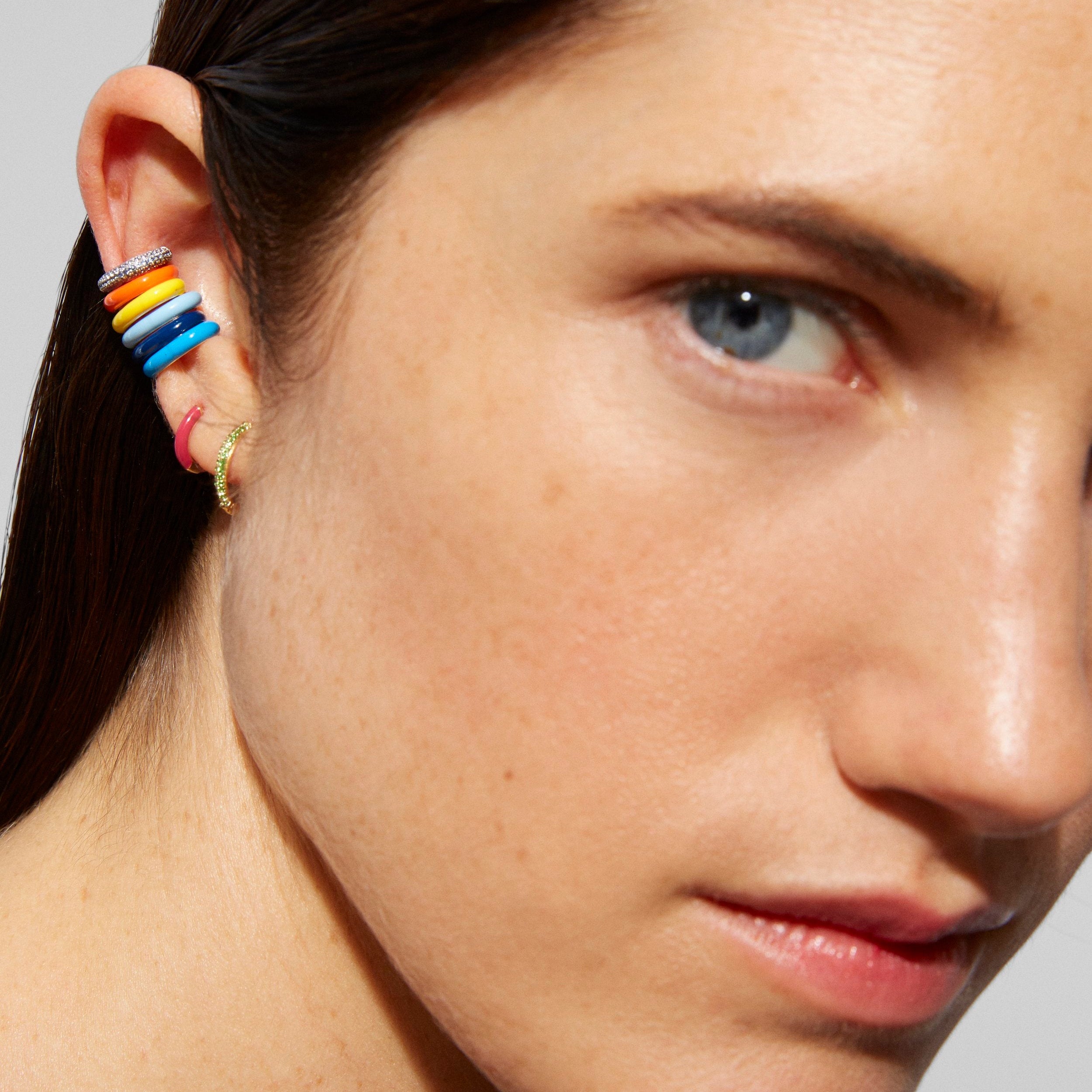 Close-up of a model's ear wearing a stack of colorful ear cuffs, including rainbow stripes and a gold hoop, against a neutral gray background.