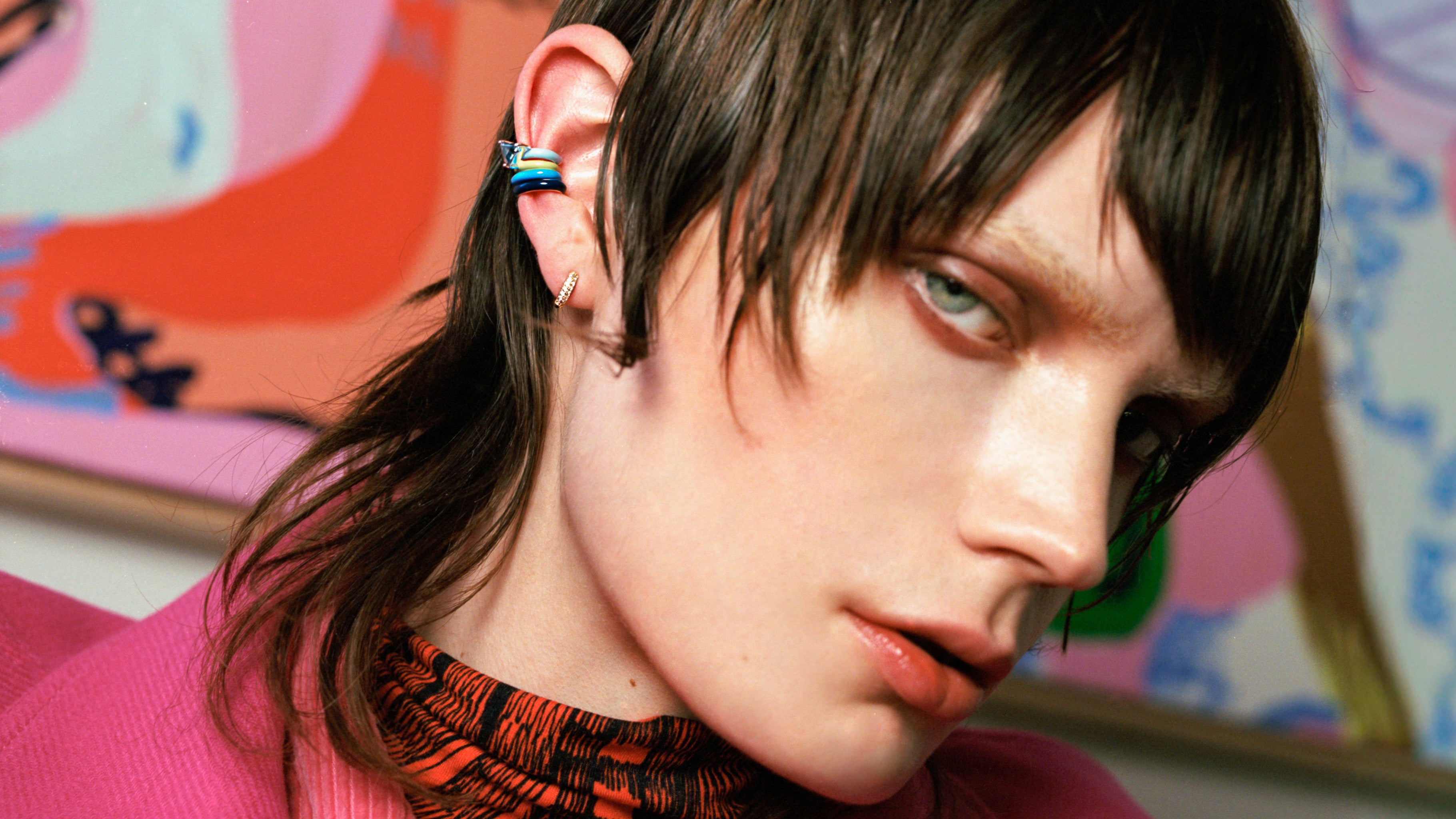 A close-up portrait of a person with dark hair wearing a colorful striped earring, a pink top, and a patterned orange neck gaiter, set against a colorful abstract background.