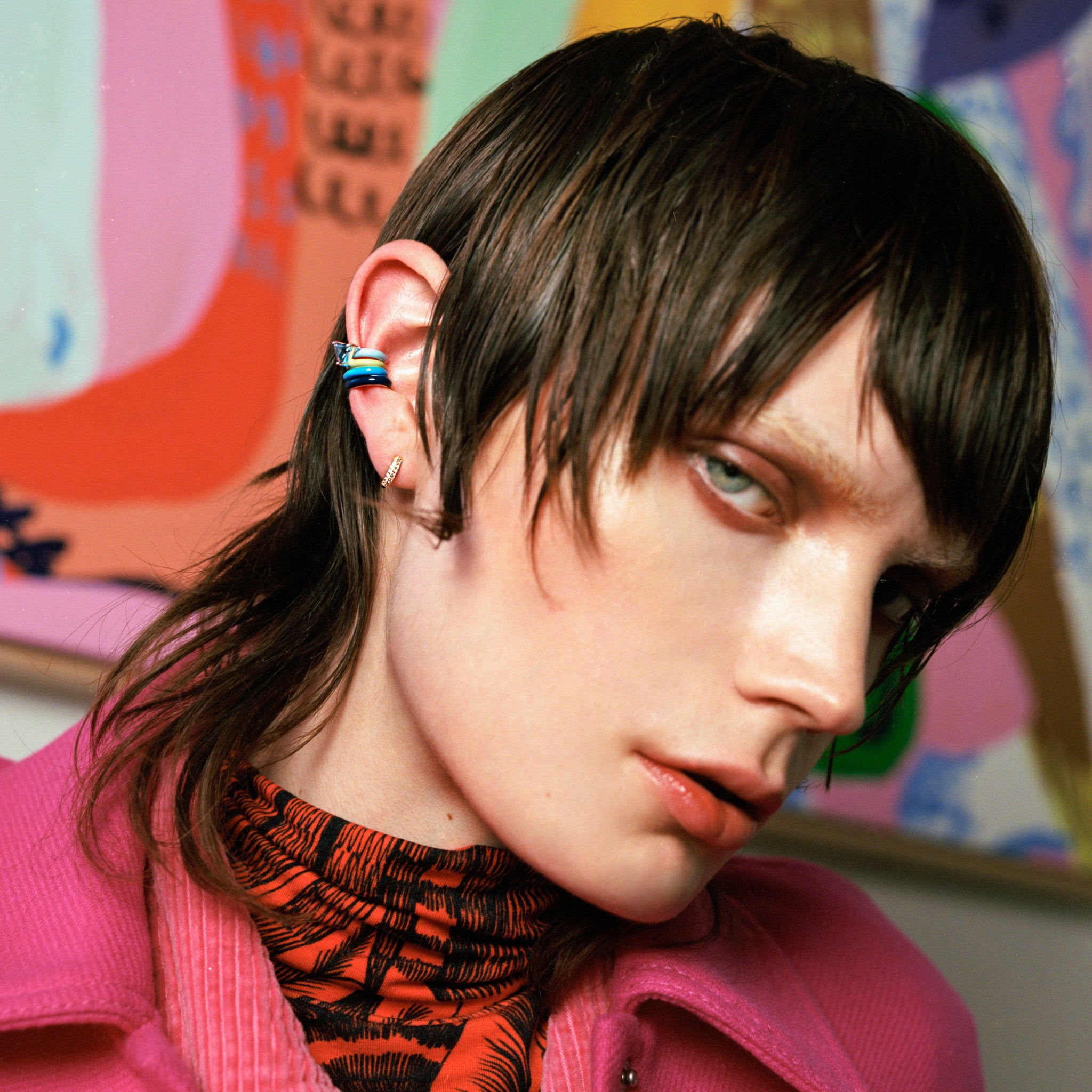A close-up portrait of a person with dark hair wearing a bright pink jacket and a black-and-orange patterned top, with colorful abstract artwork in the background.