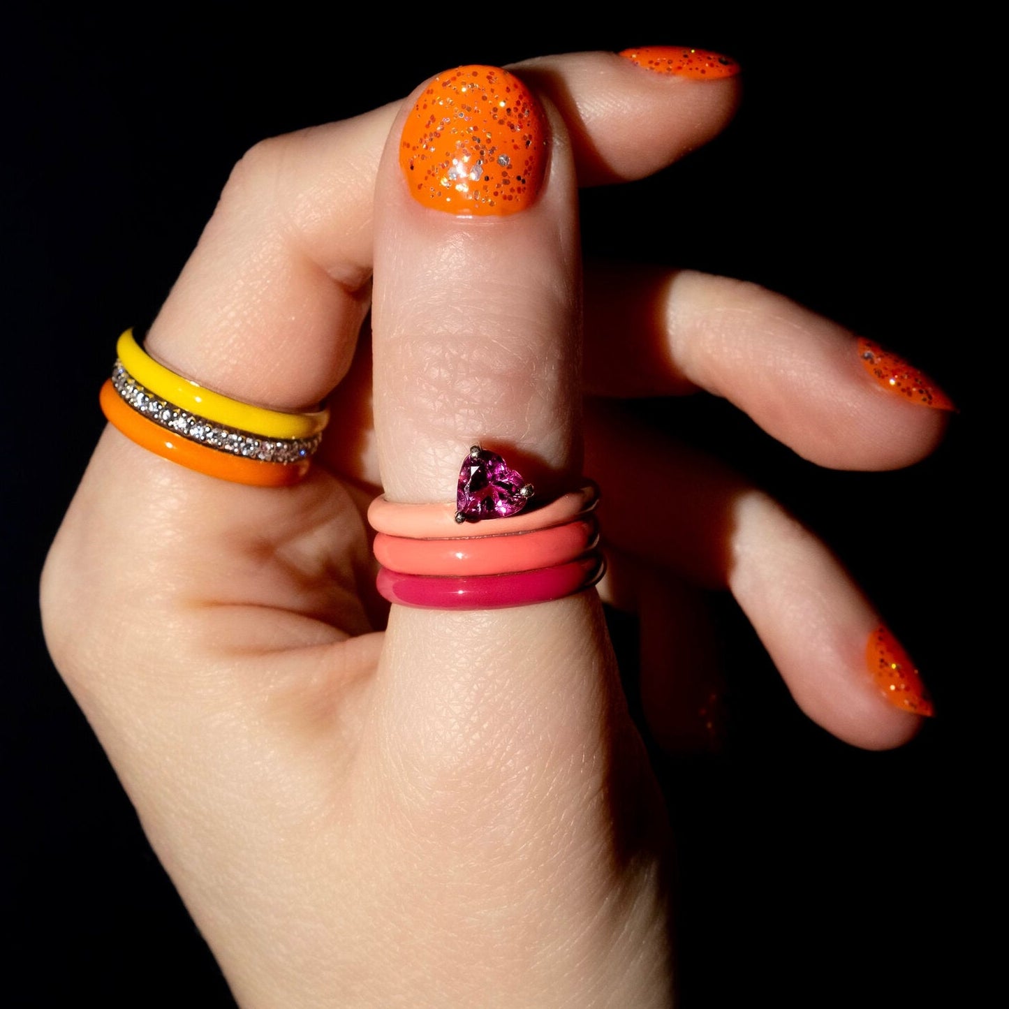 A close-up of a hand wearing multiple colorful rings against a black background. The hand has bright orange glitter nail polish, and the rings include yellow and silver bands, an orange band, a coral pink band, a magenta pink band, and a purple marquise-cut gemstone ring.