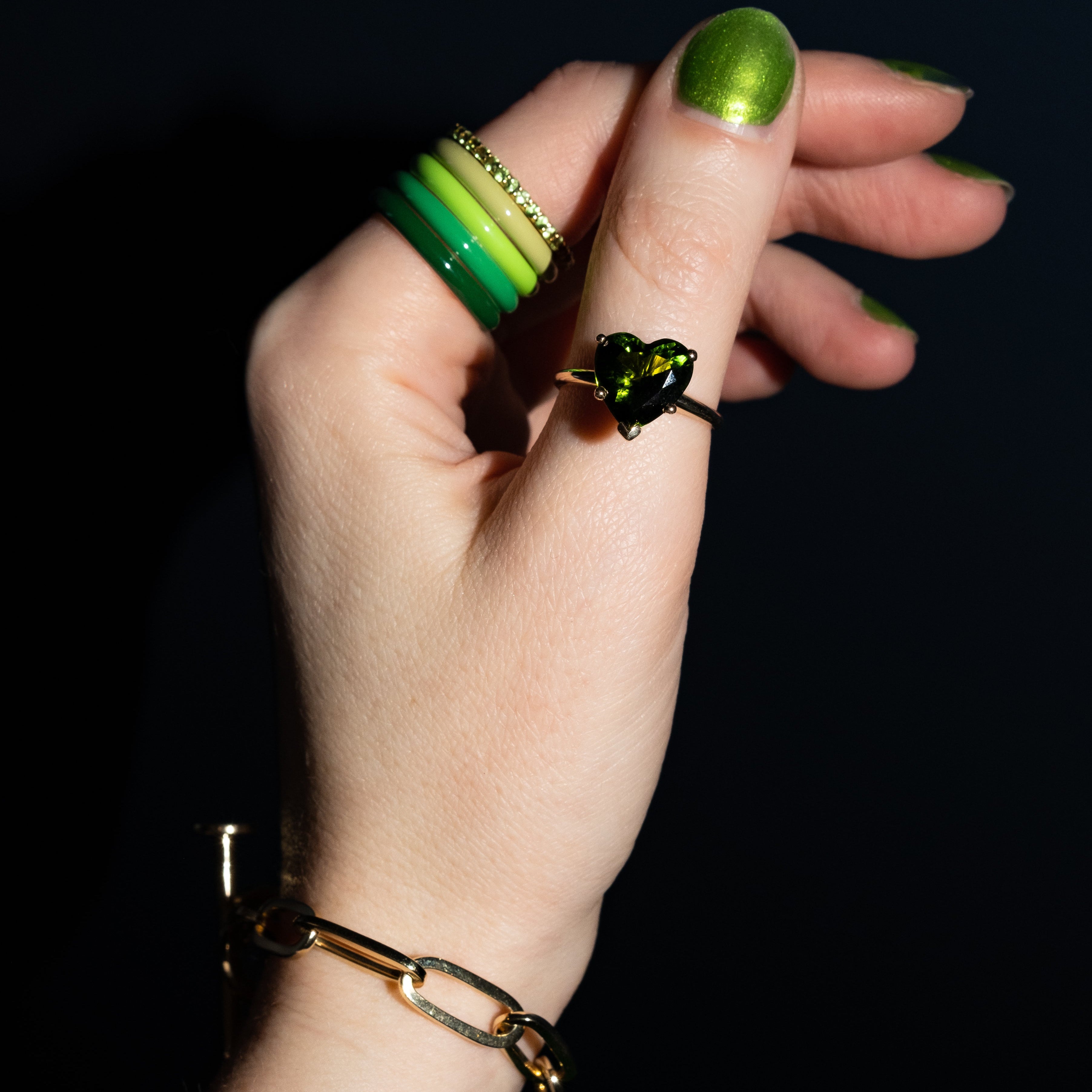 A close-up of a hand wearing multiple rings and a bracelet against a black background. The most prominent product appears to be a gold ring with a dark green heart-shaped stone on the ring finger, with additional green and yellow rings stacked on the other fingers and a gold chain bracelet on the wrist.