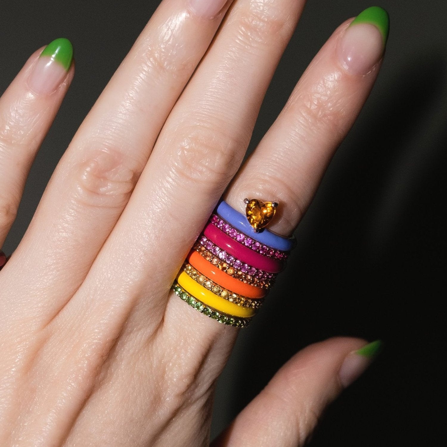 A close-up of a hand with green-tipped nails wearing a stack of colorful rings, including blue, orange, pink, yellow, and green bands with rhinestones, plus a small gold heart-shaped ring on the middle finger, against a dark background.