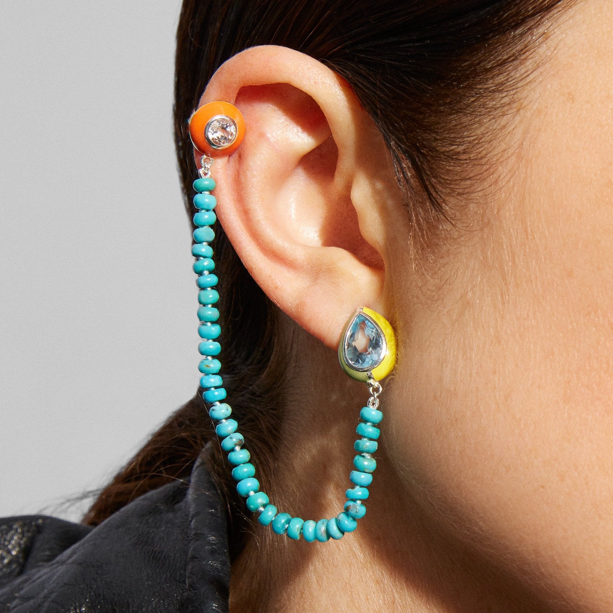 Pear-shaped topaz and enamel stud earrings with connected turquoise hand-knotted bead strand chain connects to a clip-on earring at the top of the ear showing white topaz and Florida orange enamel.