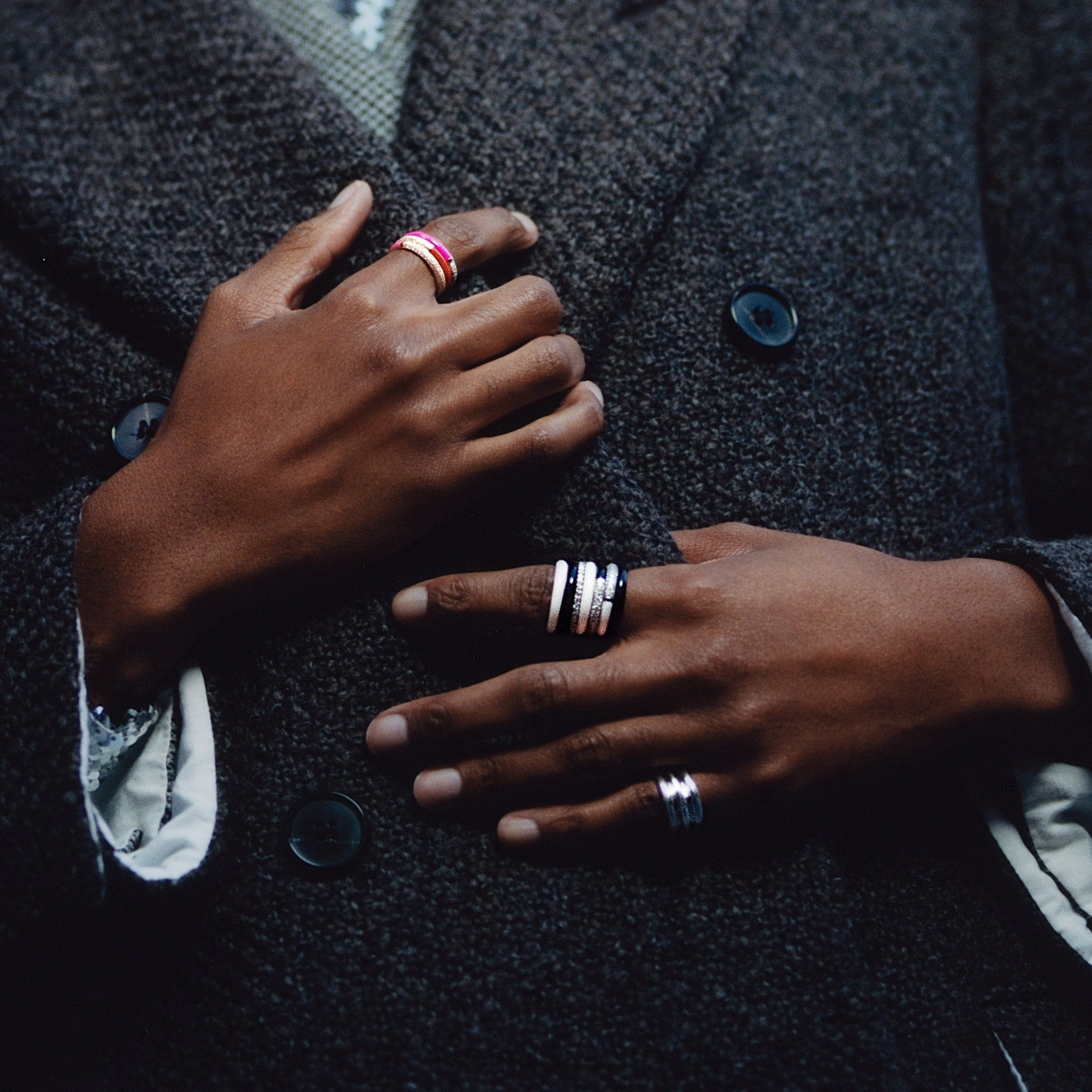 Close-up of a person wearing multiple rings on both hands, including silver band rings and a pink ring, styled with a dark textured double-breasted blazer.