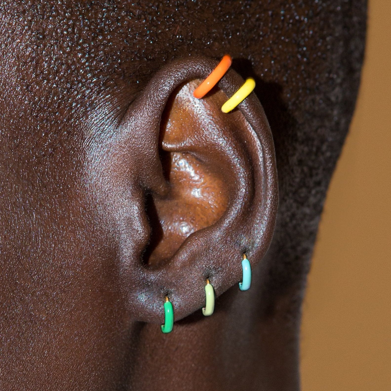 Close-up of a person's ear adorned with multiple small colorful hoop earrings in orange, yellow, green, light green, and light blue, set against dark skin and a warm tan background.