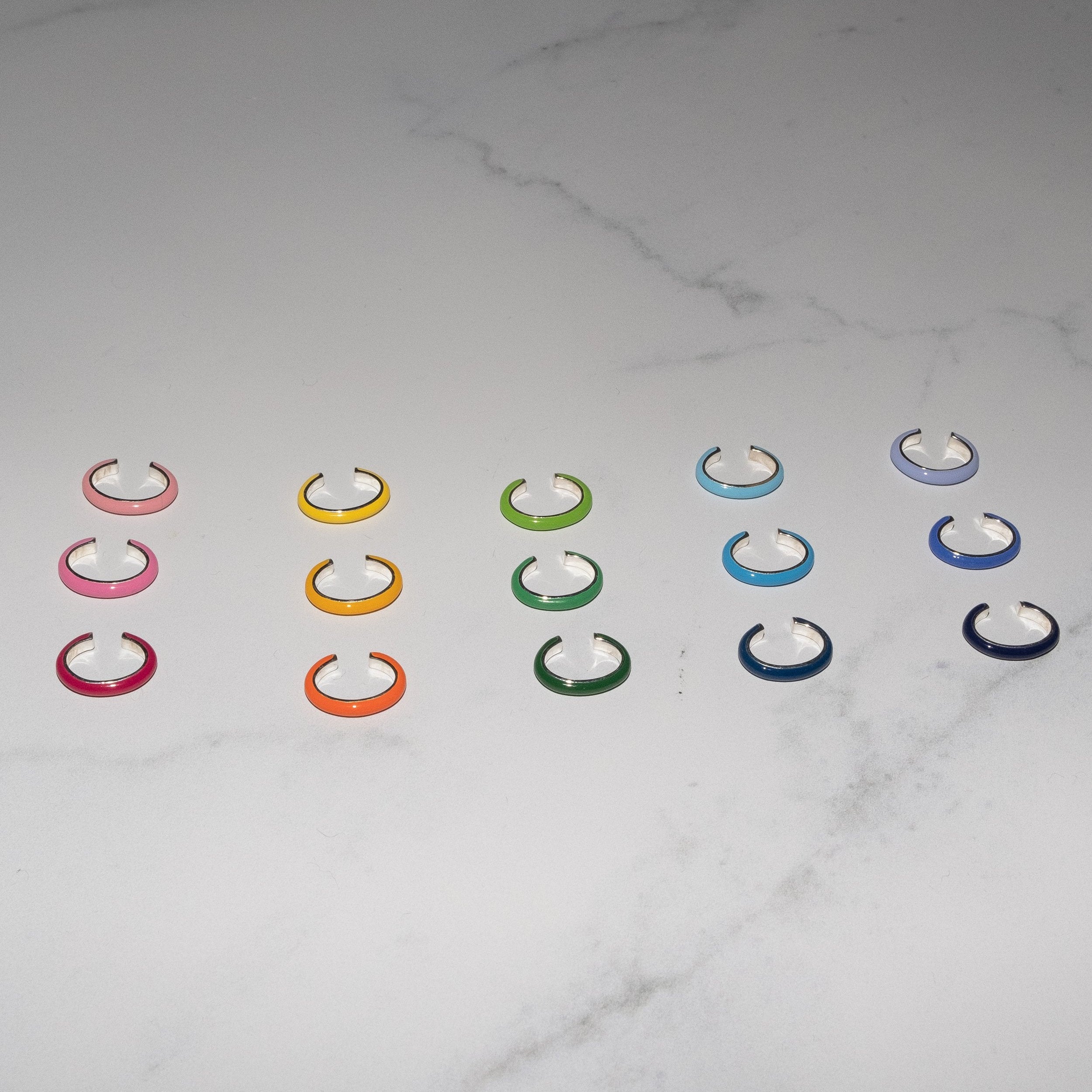 Six rows of small C-shaped ear cuff earrings in assorted colors, including pink, yellow, green, blue, orange, and purple, arranged on a white marble surface.