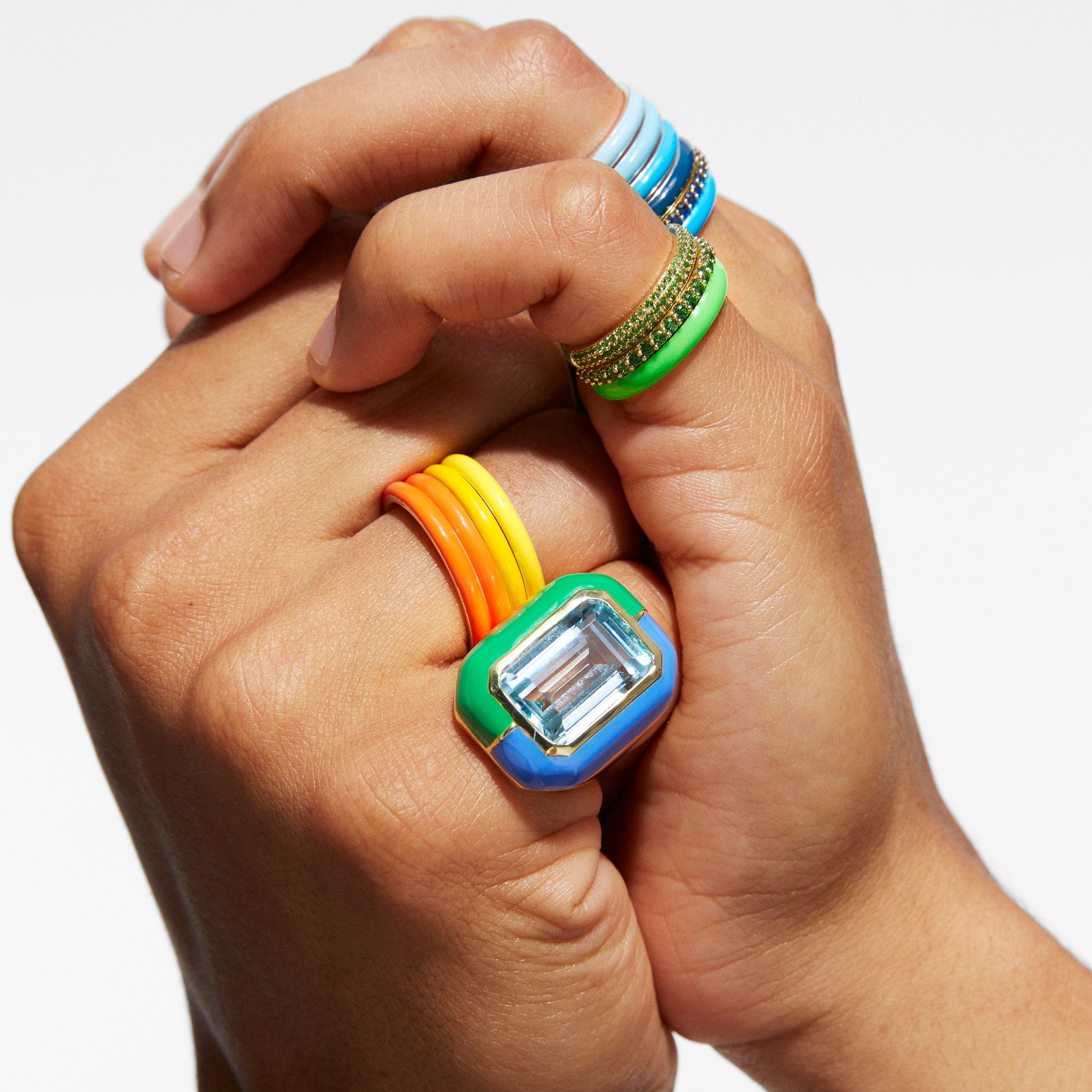A close-up of a person's hands wearing multiple colorful rings, including a prominent square ring with blue, green, orange, and yellow bands and a clear rectangular center stone.