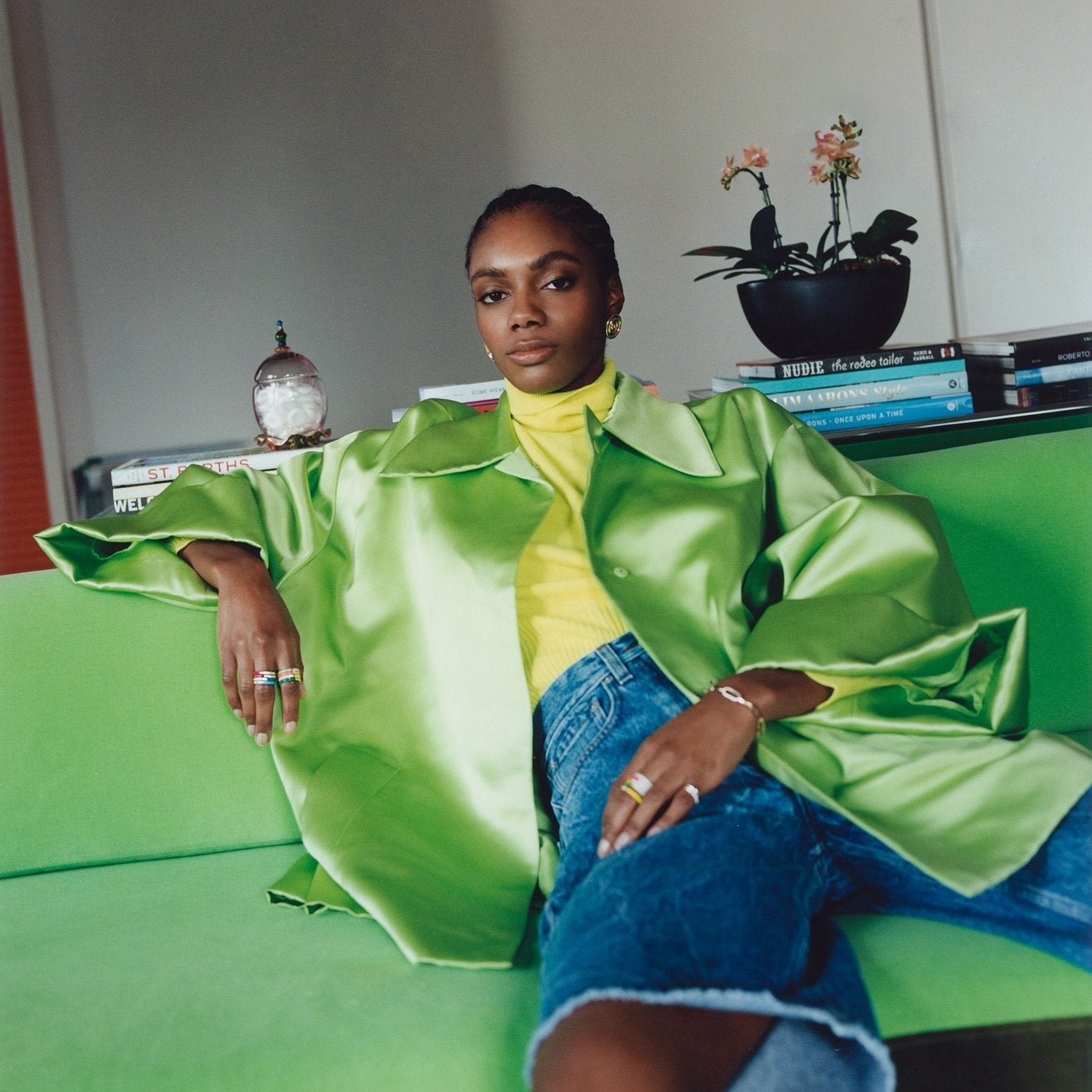 Person wearing a bright green jacket sitting on a green couch in a room with books and a plant.