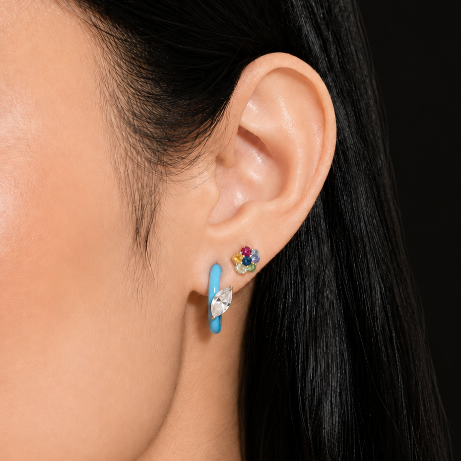 A close-up of a model's ear wearing a pair of small stud earrings, including a turquoise blue huggie hoop and a gold flower-shaped stud with colorful gemstones, against a dark background.
