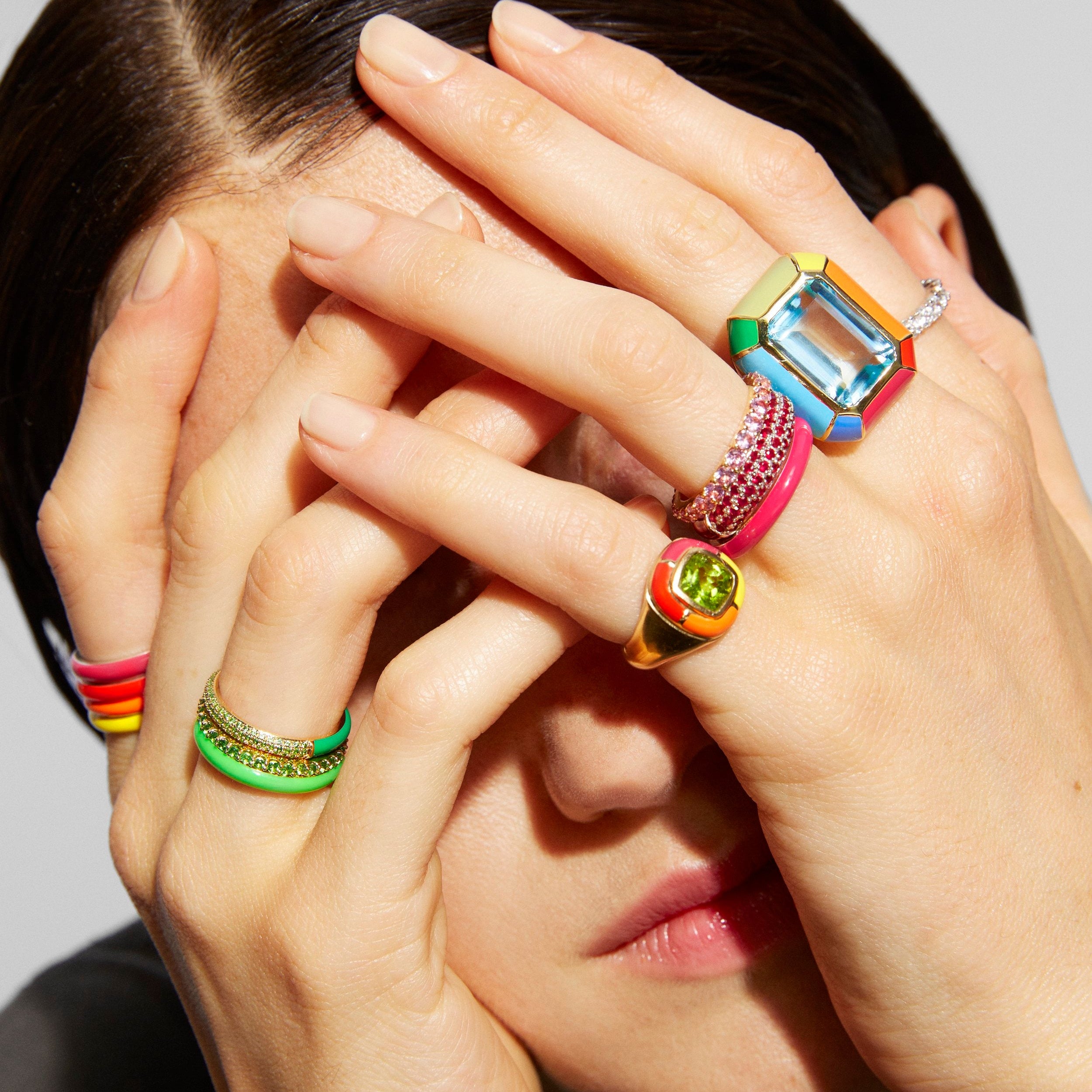 A close-up lifestyle shot of a model wearing multiple colorful rings. The rings feature vibrant colors like green, pink, yellow, orange, and blue, with one ring showing a clear rectangular gemstone and another featuring a gold-toned signet-style face.