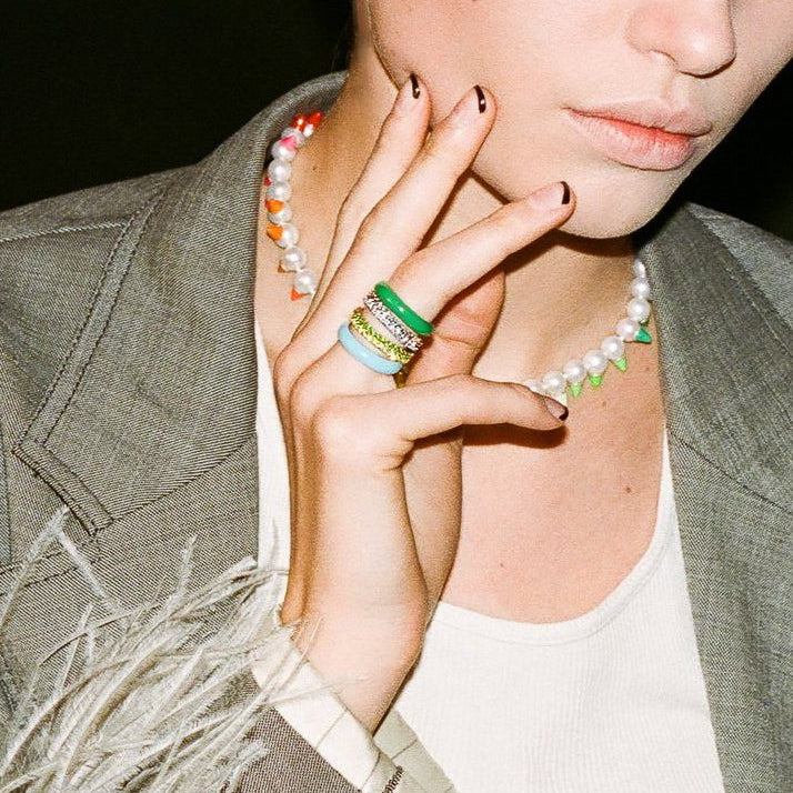 A close-up lifestyle shot of a model wearing a stack of colorful rings, including a green and white striped ring, a light blue ring with a crystal band, and a white ring with a black crystal band. The model is also wearing a pearl necklace and a gray blazer.