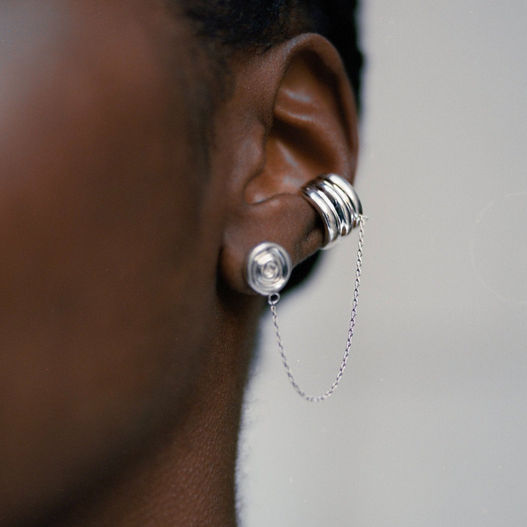 A close-up side profile of a person's ear wearing a silver hoop earring with a long dangling chain and stacked silver hoop details behind the ear.