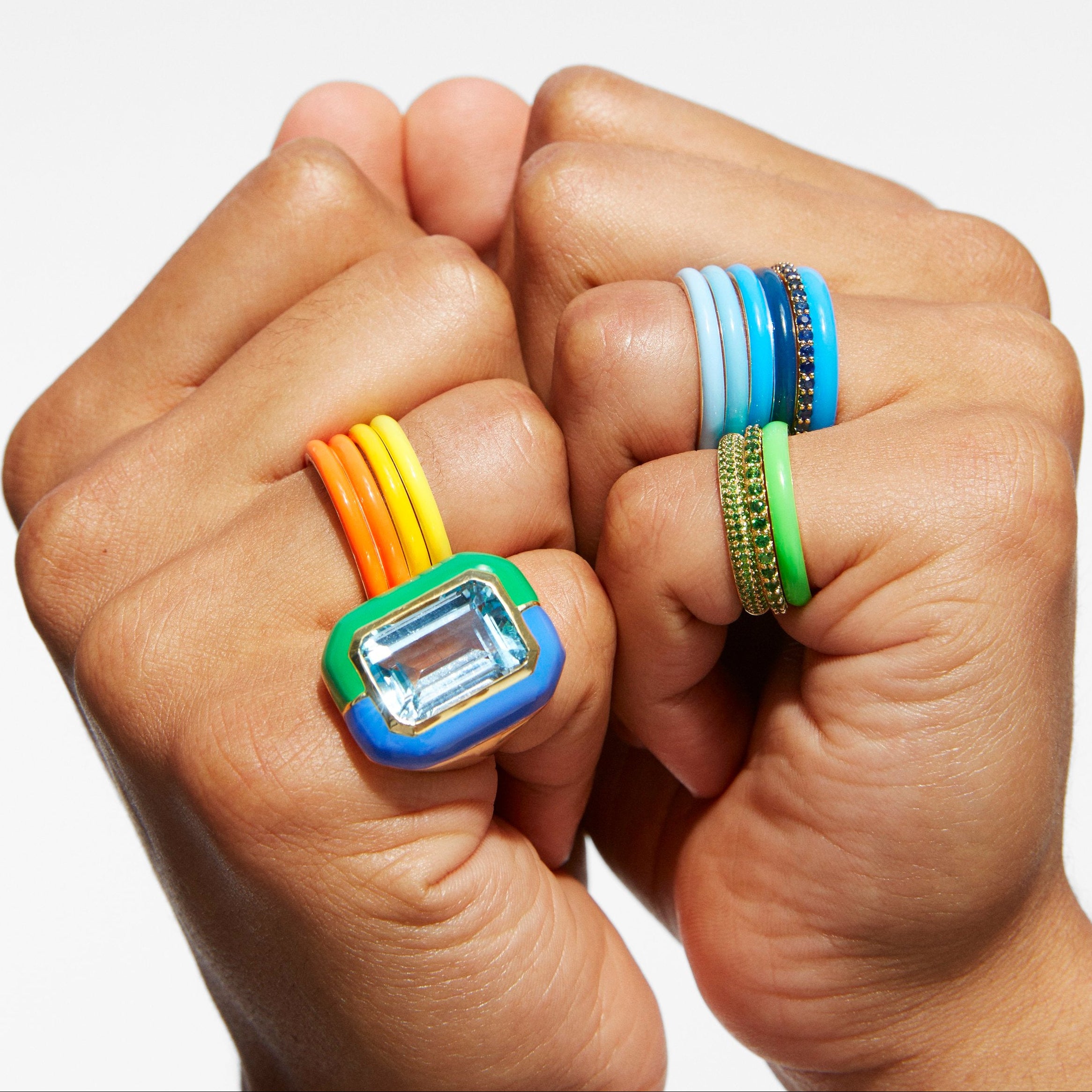 Close-up of two hands wearing colorful stretch rings against a light gray background. One ring has a blue rectangular face with a clear gemstone and orange and yellow elastic bands.