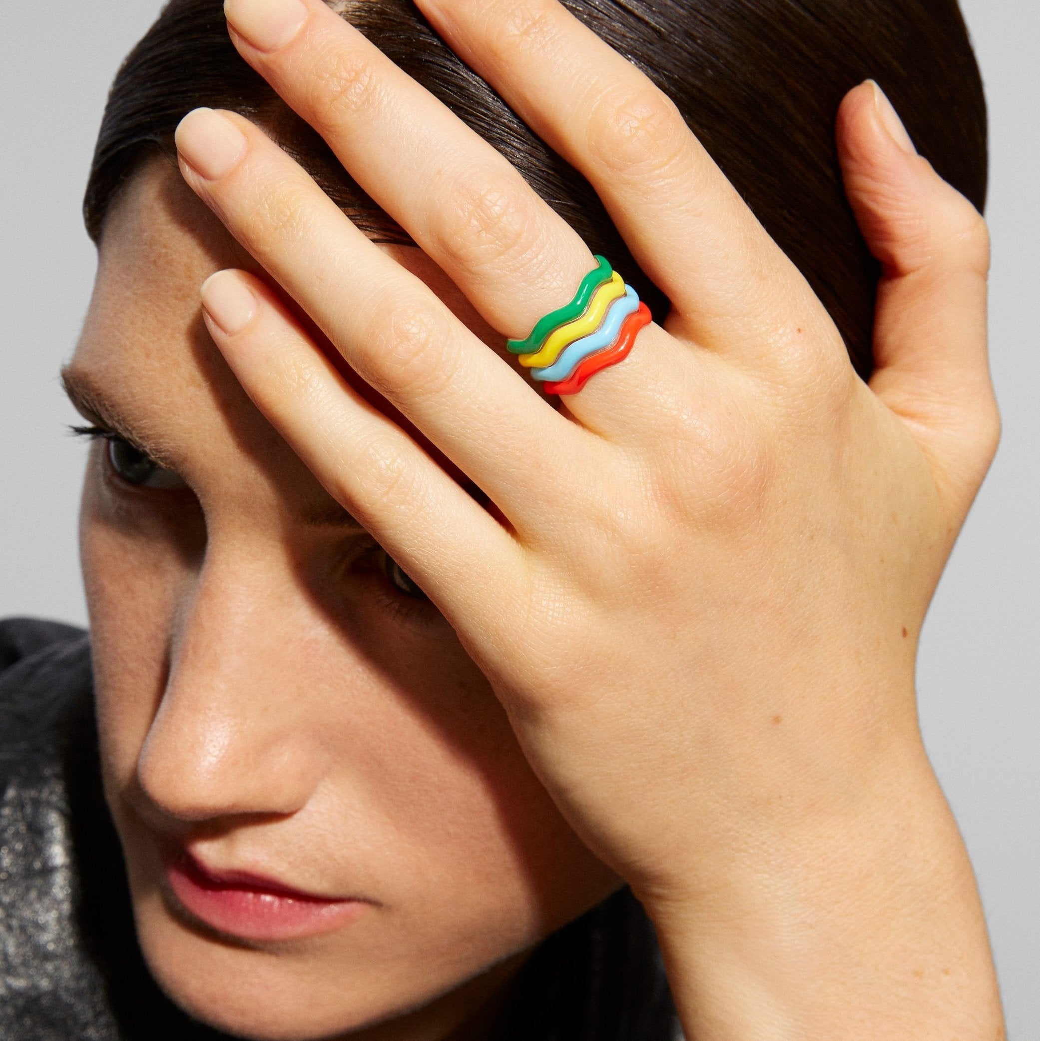 A close-up lifestyle shot of a model wearing a wavy, multi-colored ring on her finger, with her hand covering part of her face against a neutral gray background.