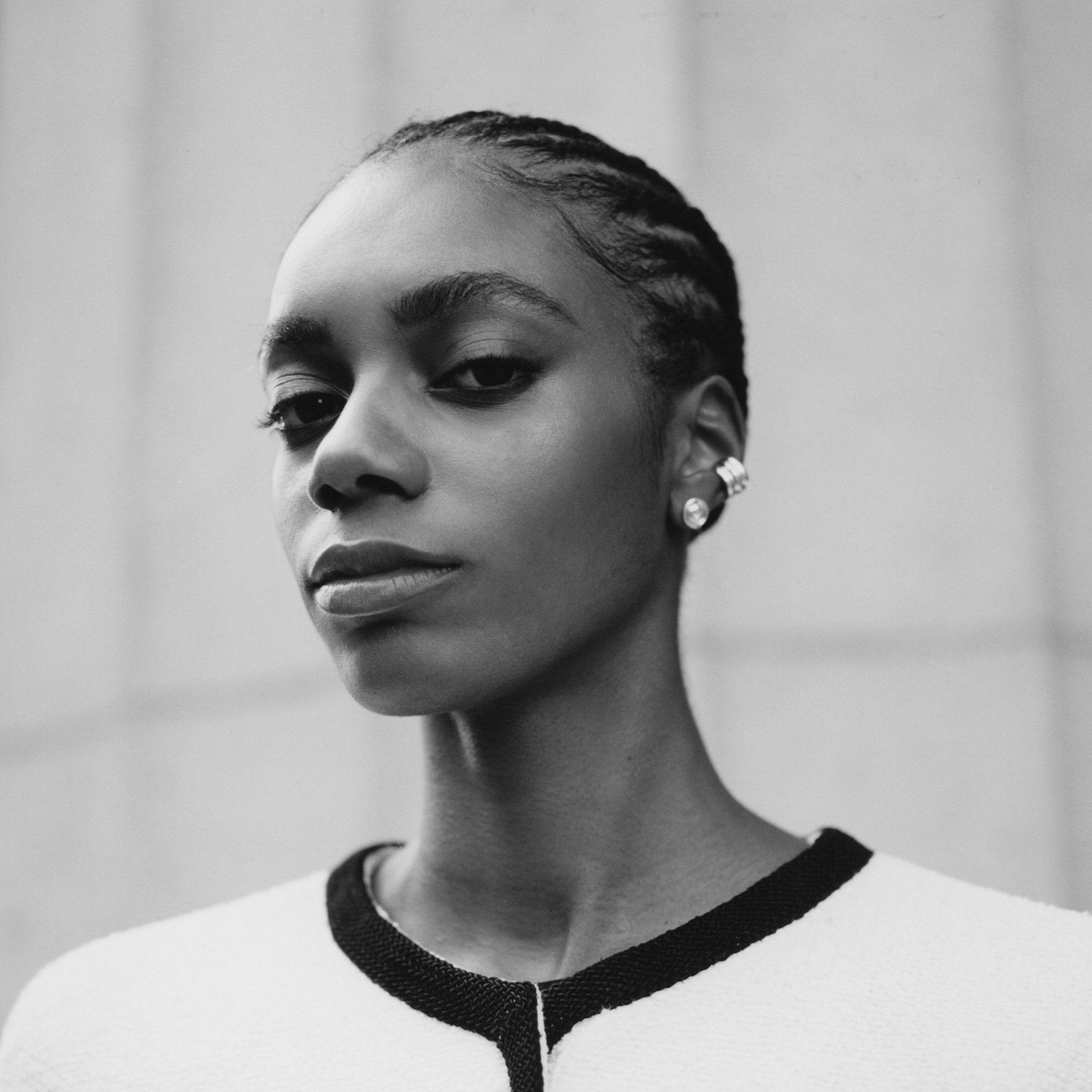 A black and white portrait of a woman wearing a white textured jacket with black trim and pocket details, set against a neutral stone wall background.