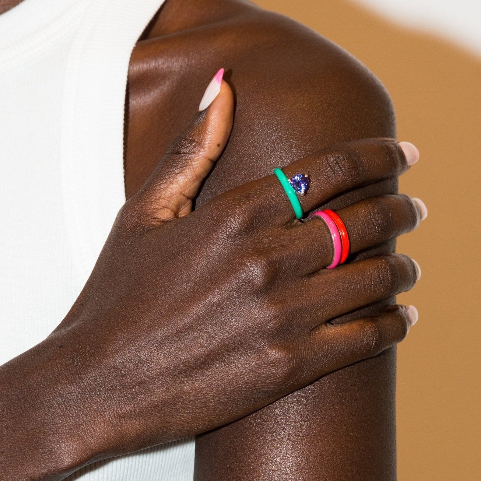 Model's hand wearing a fuchsia ring with a red ring and green ring with blue gemstone heart grouped on the hand.