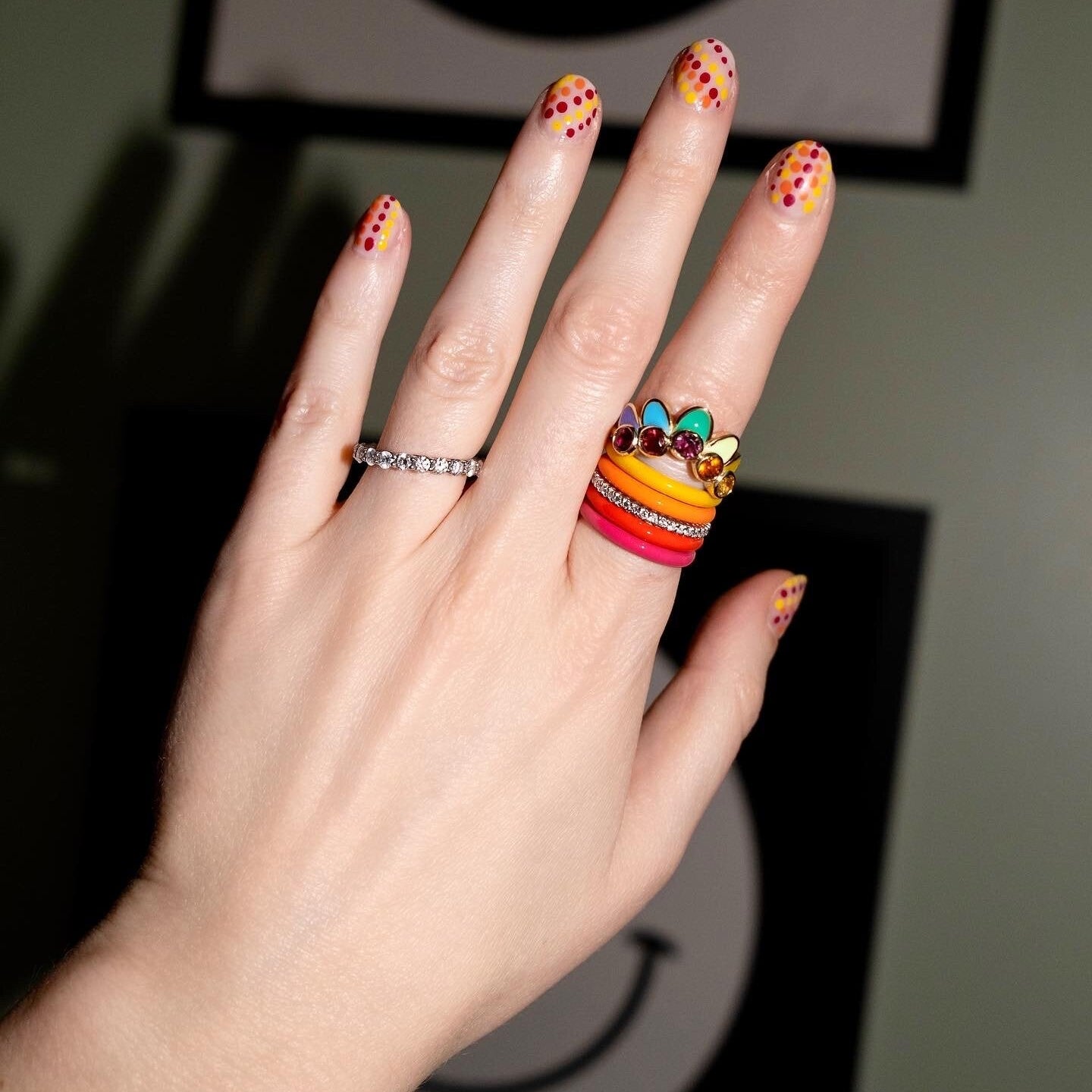 A close-up of a hand wearing a stack of colorful rings, with the most prominent ring featuring yellow, orange, and red bands and a small multicolor jewel cluster on top. The hand has yellow and red polka-dot manicured nails, and the background is a dark, softly blurred interior setting.