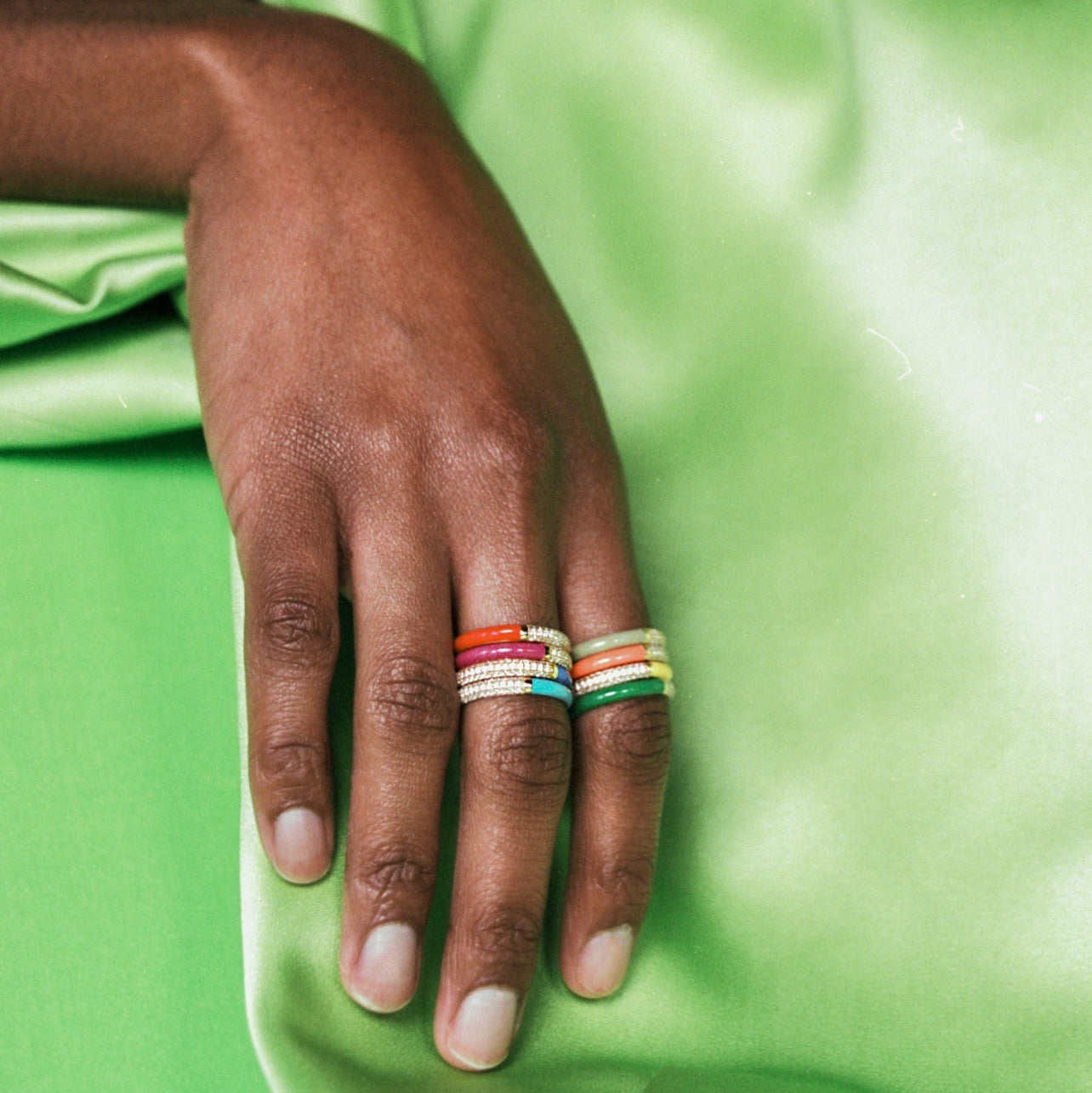 A close-up of a hand resting on a bright green satin fabric, wearing two stacked rings made of colorful woven threads.