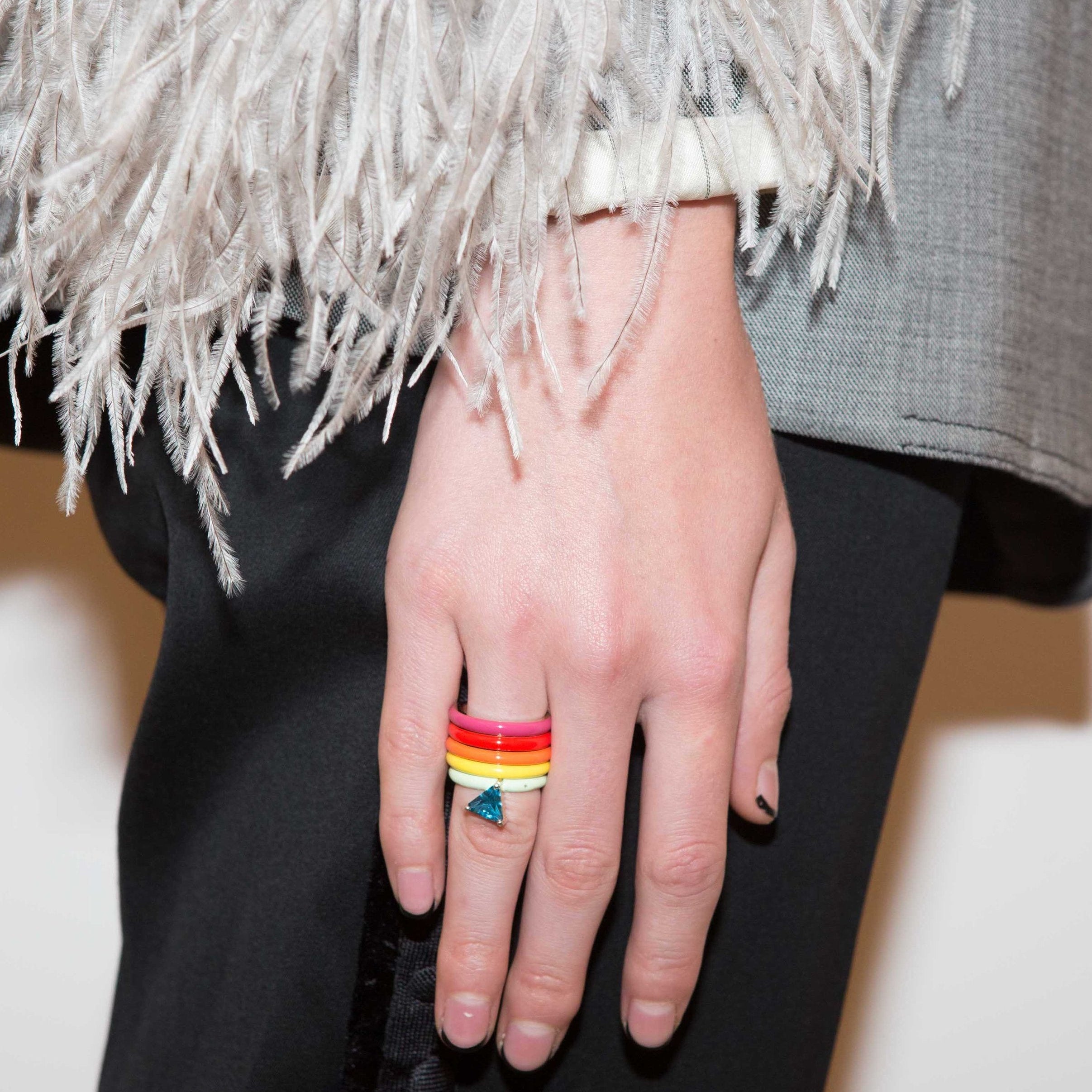 A close-up of a person's hand wearing a stack of colorful rings, including a red, orange, and yellow striped band and a small blue triangular ring, styled with a gray jacket featuring white feathers and black trousers.