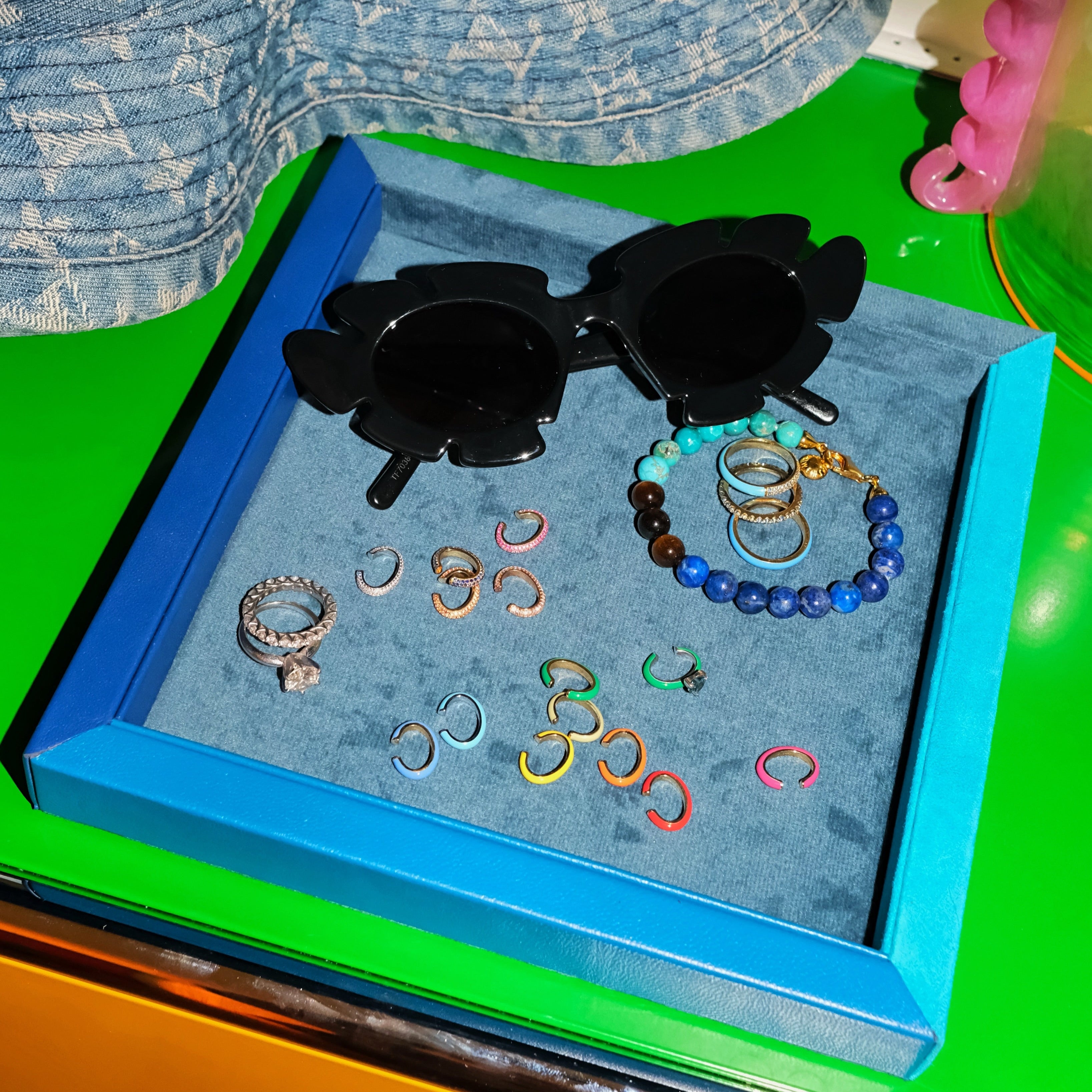 A collection of fashion accessories displayed on a blue velvet jewelry tray, including black flower-shaped sunglasses, a beaded bracelet, and multiple rings, set against a bright green surface.