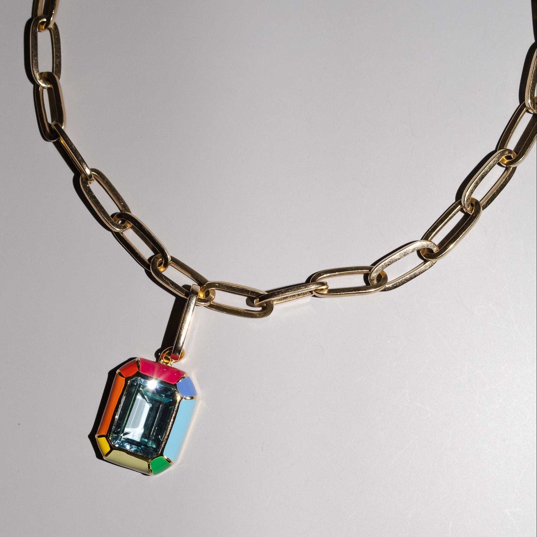 A gold-toned paperclip chain necklace with a colorful faceted gemstone pendant, displayed on a neutral gray background.