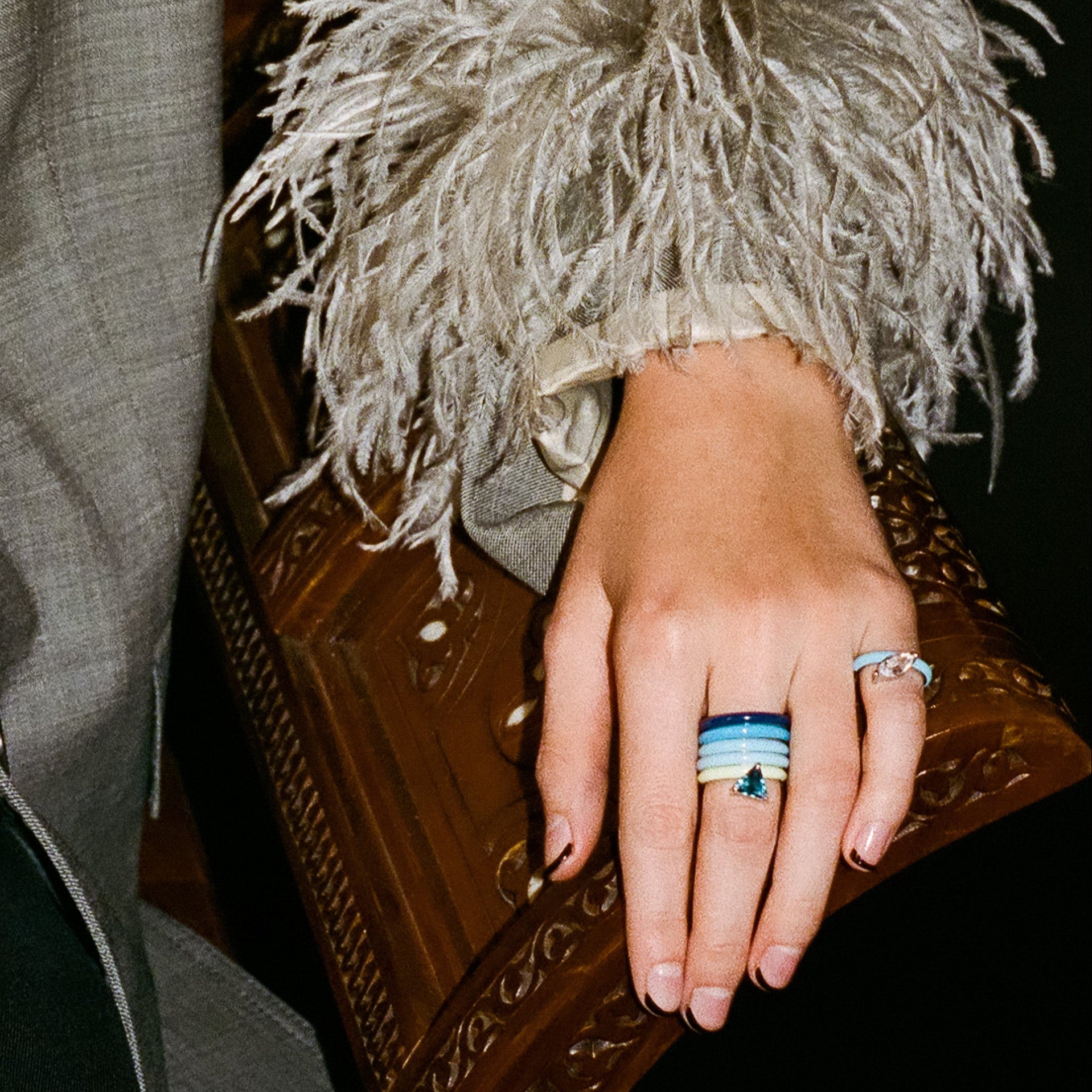 A close-up of a person's hand resting on an ornate brown wooden box, wearing a silver ring with a blue and white striped stone and another silver band. The person is wearing a gray textured jacket with a large, fluffy light gray feathered cuff.