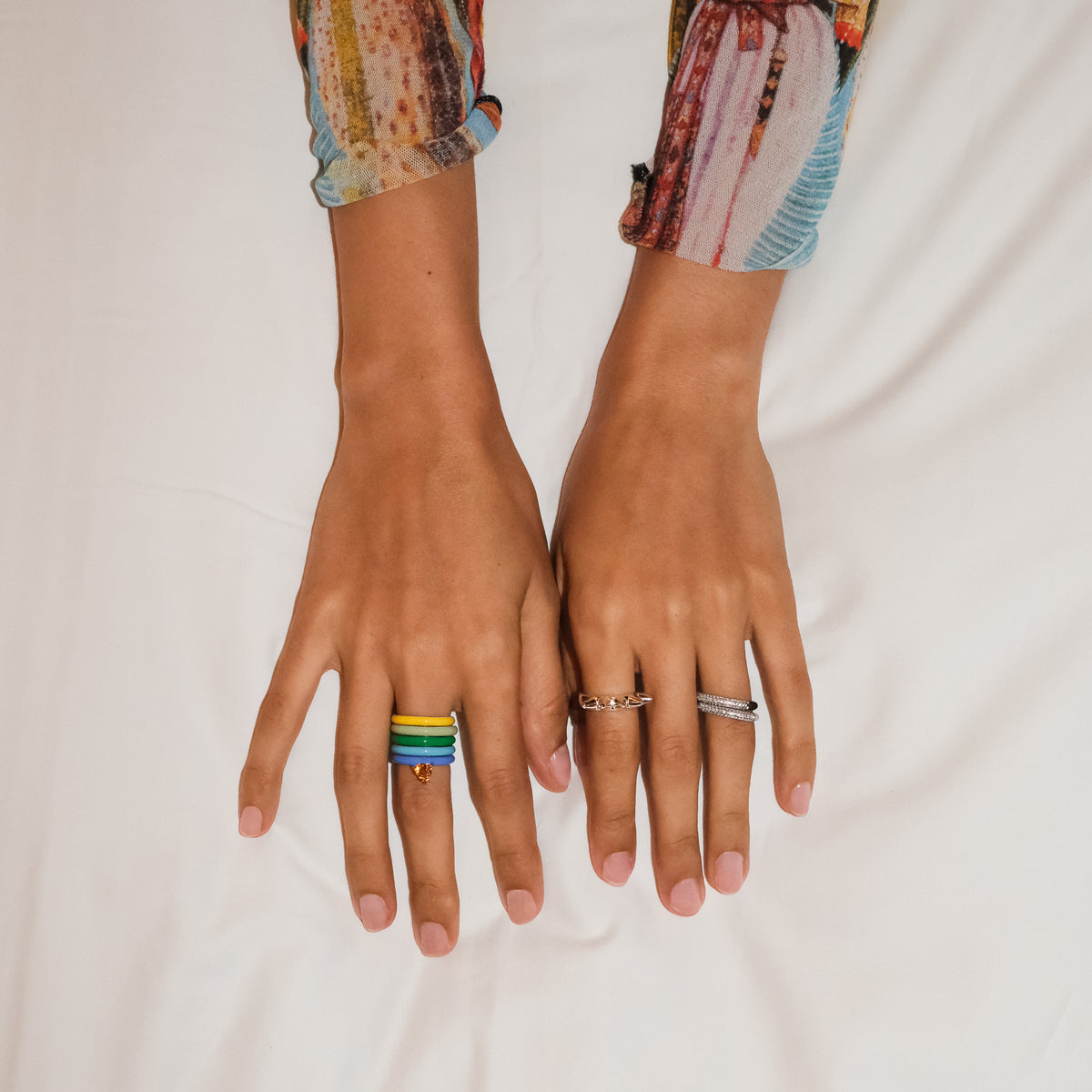 A top-down view of a person's hands resting on white fabric, showcasing a colorful-striped set of five rings on the left hand featuring an orange citrine heart gemstone and two silver rings on the right hand. The person is wearing a long-sleeve shirt with a multicolor abstract pattern.