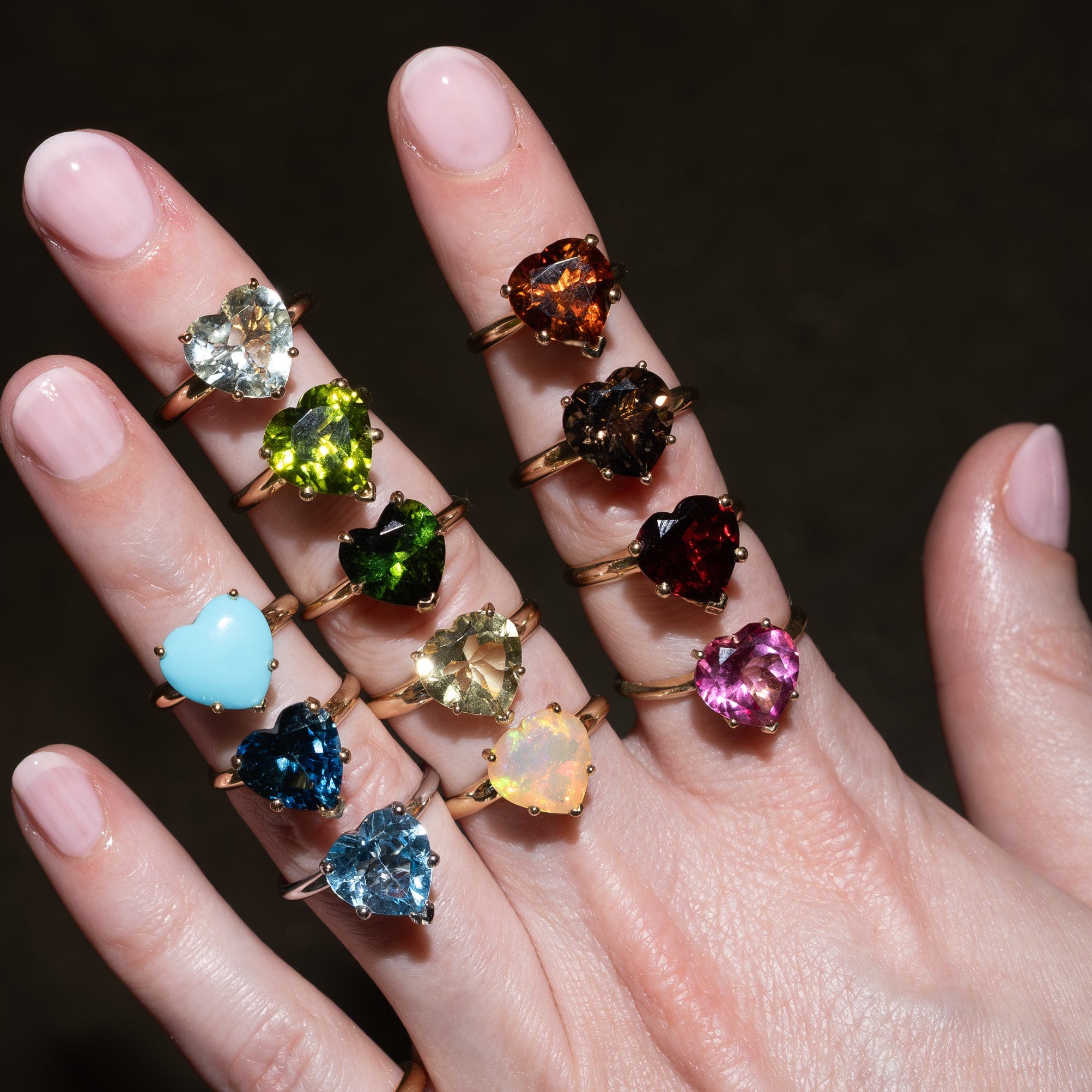 A hand with pale pink manicured nails wearing multiple gold rings, each featuring a different colored heart-shaped gemstone, set against a dark background.