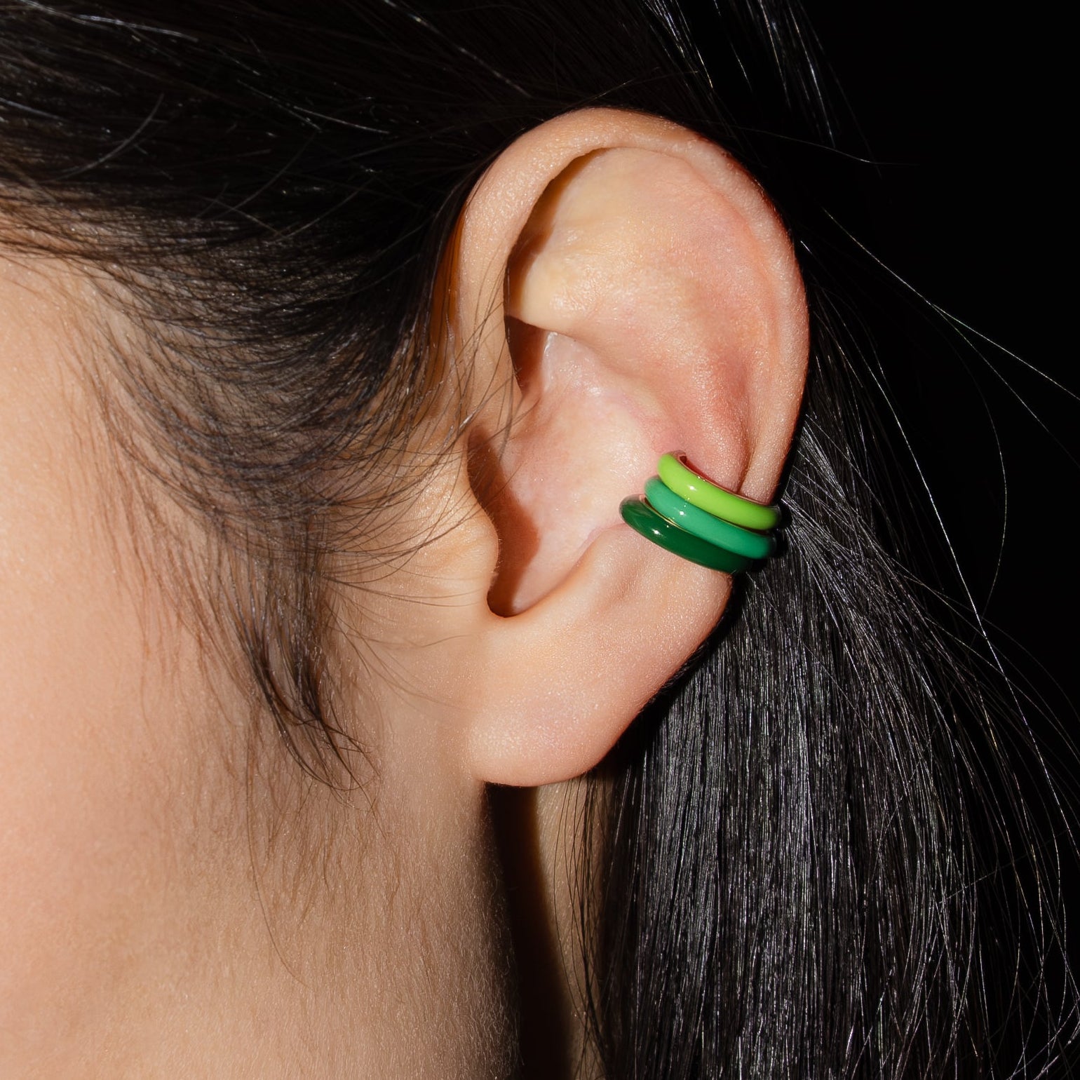 Close-up of an ear with three green ear cuff earrings against a dark background.