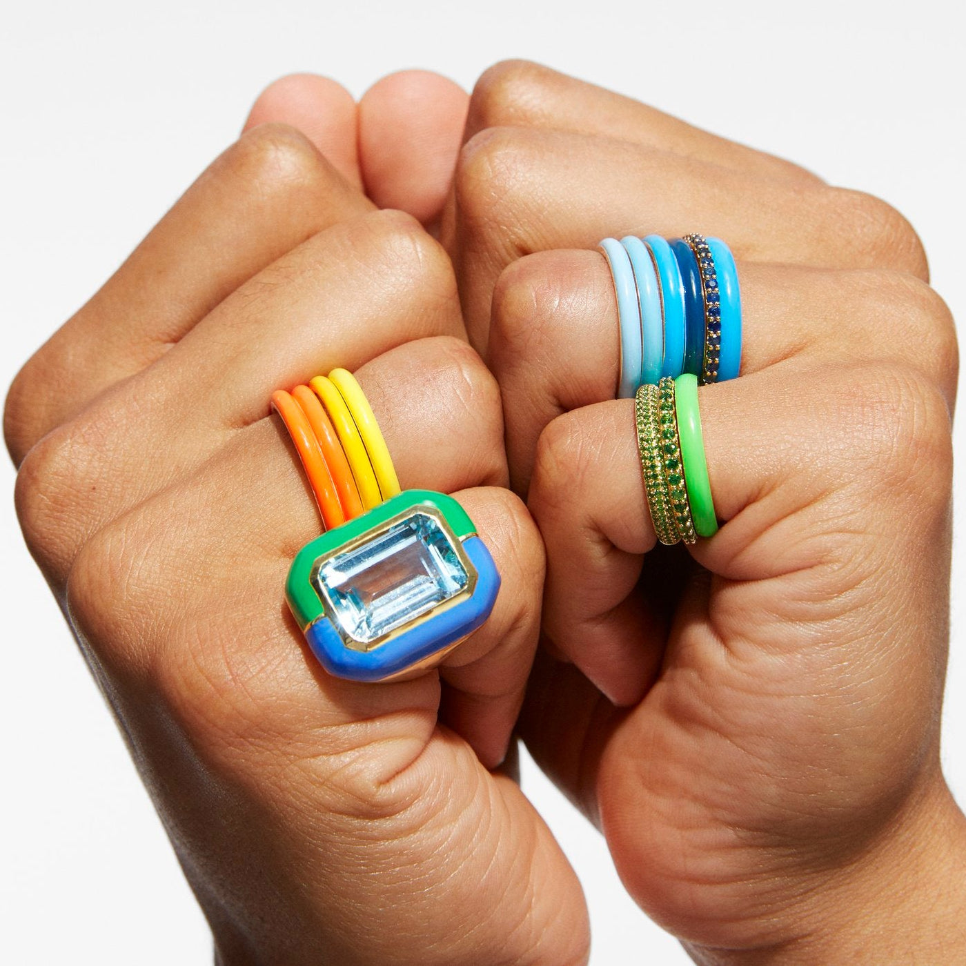 Close-up of two hands wearing colorful rings against a light gray background. One ring features a large clear rectangular gemstone with blue and green settings and orange and yellow bands.