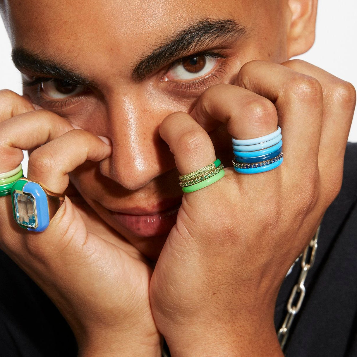 Close-up of a person wearing multiple colorful rings, including stacked blue and light blue bands, a green band with gold detailing, and a large blue and clear stone ring, against a light background.