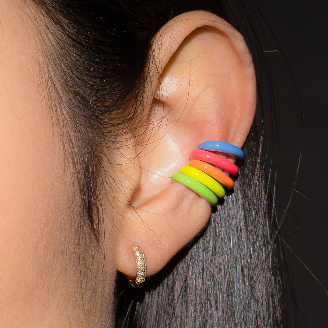Close-up of an ear with a stack of five rainbow-colored ear cuff earrings against a dark background.