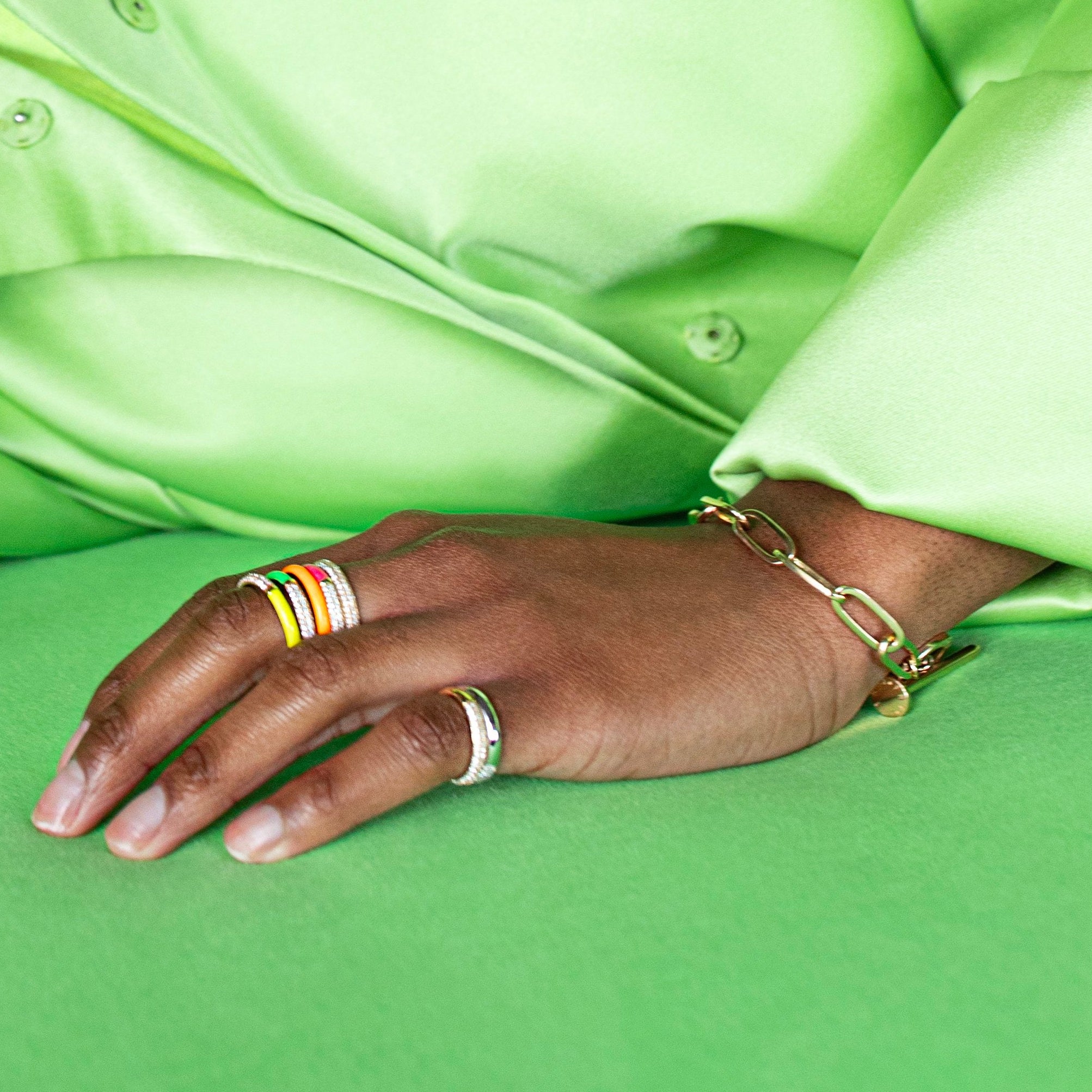 A close-up of a person's hand resting on a green surface, wearing a gold chain bracelet on the wrist and multiple colorful rings on the fingers, with a bright lime green satin shirt in the background.