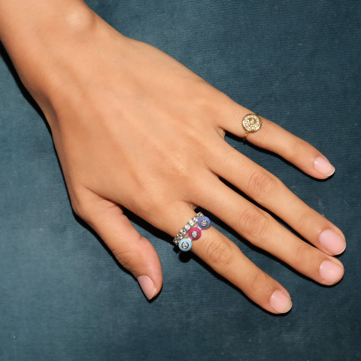 A close-up of a hand wearing two rings against a dark teal textured background. One ring is a gold signet-style ring with a circular face, and the other is a silver band with three colorful enamel bezels around white sapphire stones.