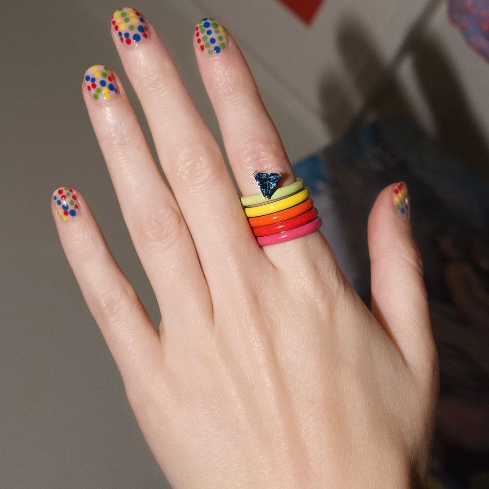 A close-up of a hand wearing a stack of five brightly colored rings in yellow, orange, red, pink, and blue, with multicolored polka dot nail art on the other fingers.