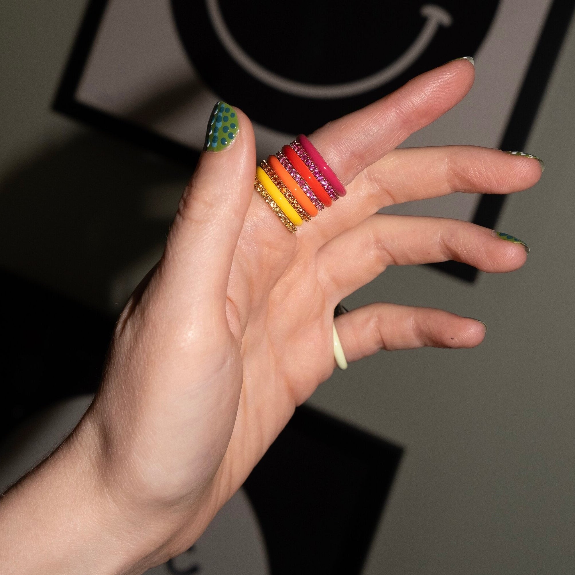 A close-up of a hand wearing five stacked colorful rings with silver-toned metal and patterned details, against a dark gray background with a framed smiley face.
