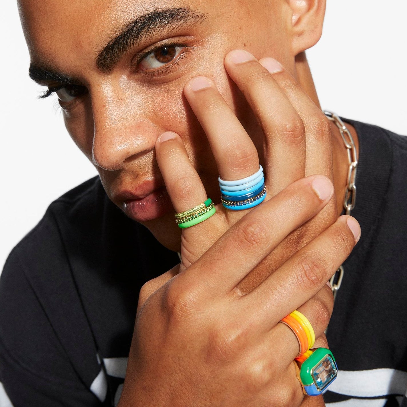 A close-up lifestyle shot of a man wearing multiple colorful rings on his fingers, including a green textured band, a blue and white striped band, and a bright orange and green watch band, against a clean white background.