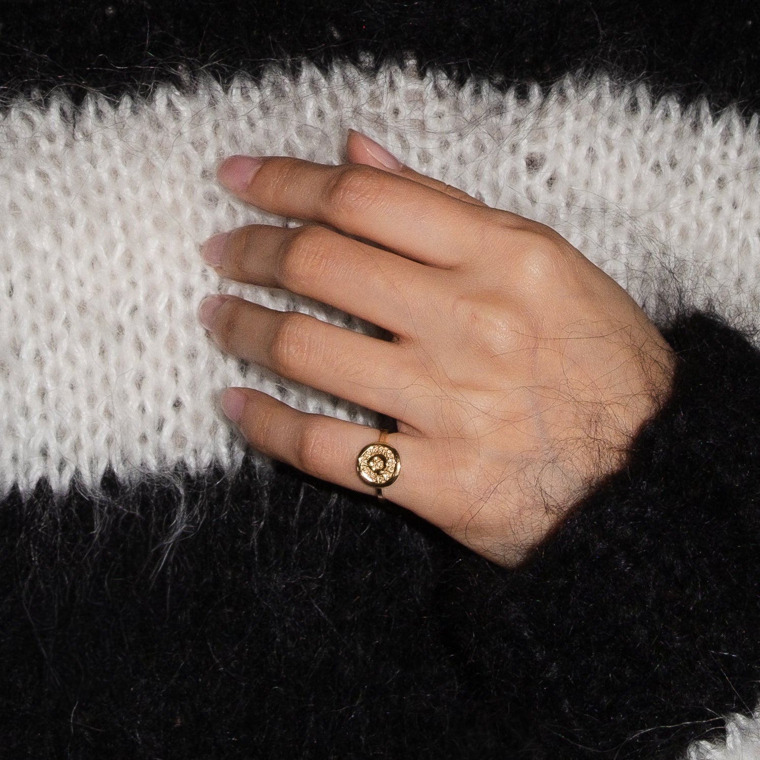 A close-up of a person wearing a fuzzy black and white striped knit sweater, with their hand resting on their chest and wearing a small gold ring.