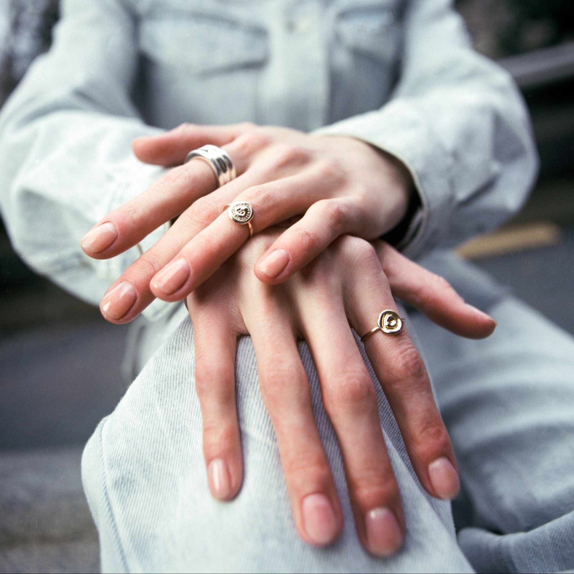 A close-up of a person's hands resting on their lap, wearing four distinct rings: a silver band with a white stone, a gold ring with a heart, a silver ring with a circular motif, and a gold ring with a heart-shaped stone. The person is wearing a light gray denim jacket and matching jeans, with a blurred outdoor background.