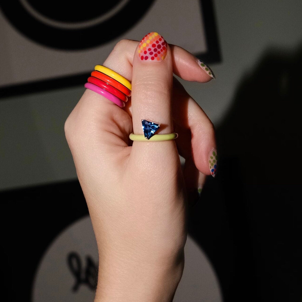 A hand wearing multiple colorful rings, with a thin yellow-green ring on the index finger featuring a small blue triangular charm. The nails are decorated with colorful patterns, and the background is dark with some abstract shapes.