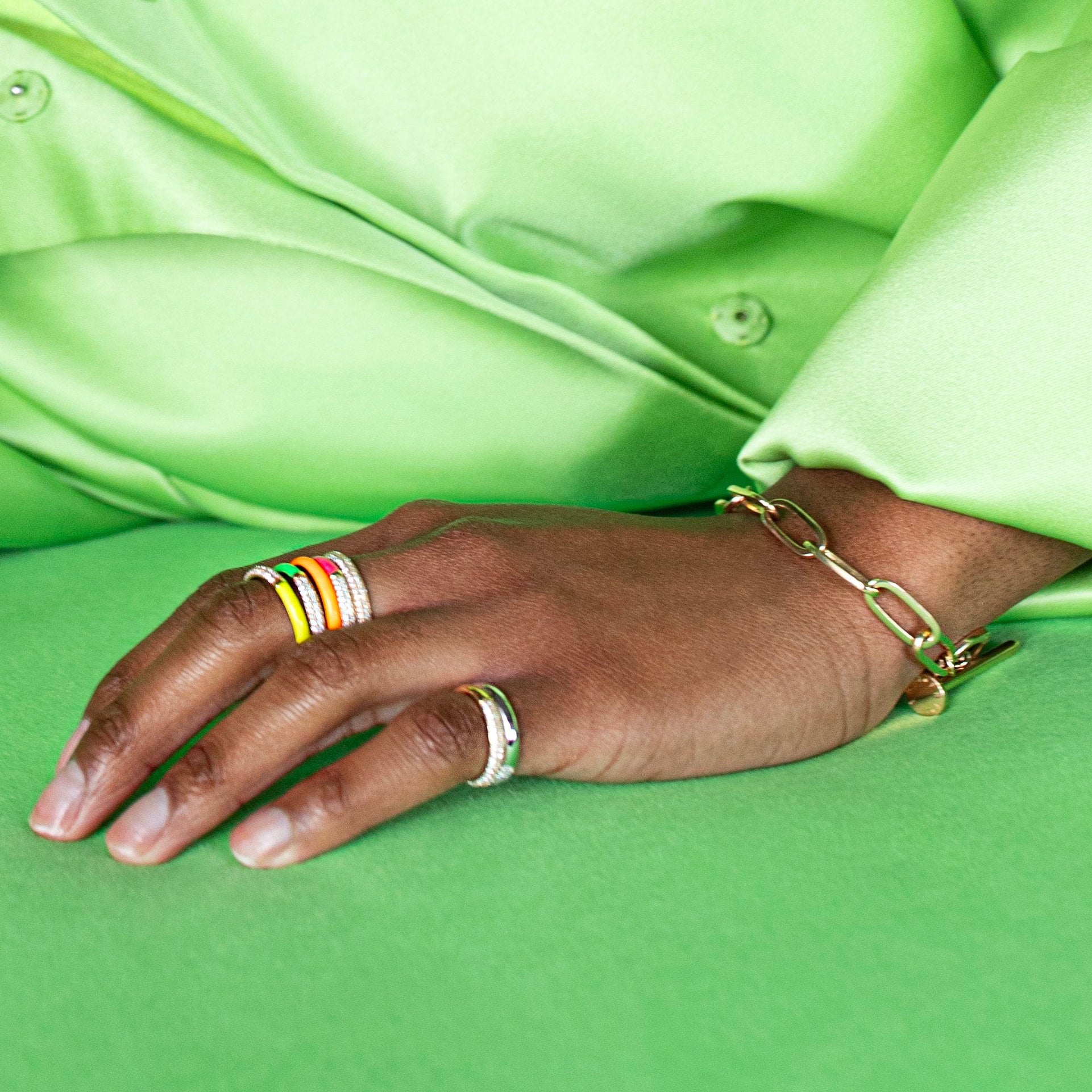 A close-up of a person's hand resting on a green surface, wearing a gold chain bracelet on the wrist and multiple colorful rings on the fingers, with a bright lime green satin shirt in the background.